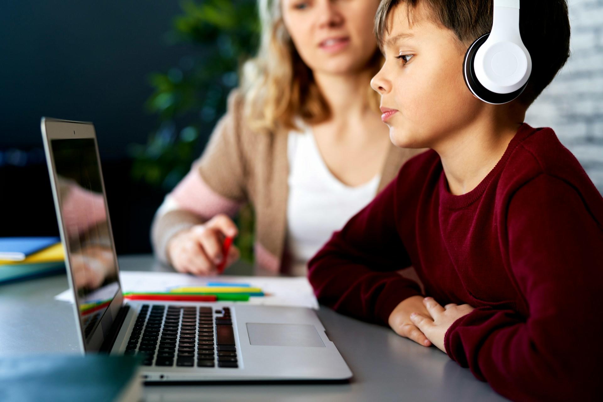 A youth wearing a dark red jumper and white headphones over his ears focuses on the open laptop computer in front of him while an adult wearing a white shirt under a tan short sleeved cardigan looks on.