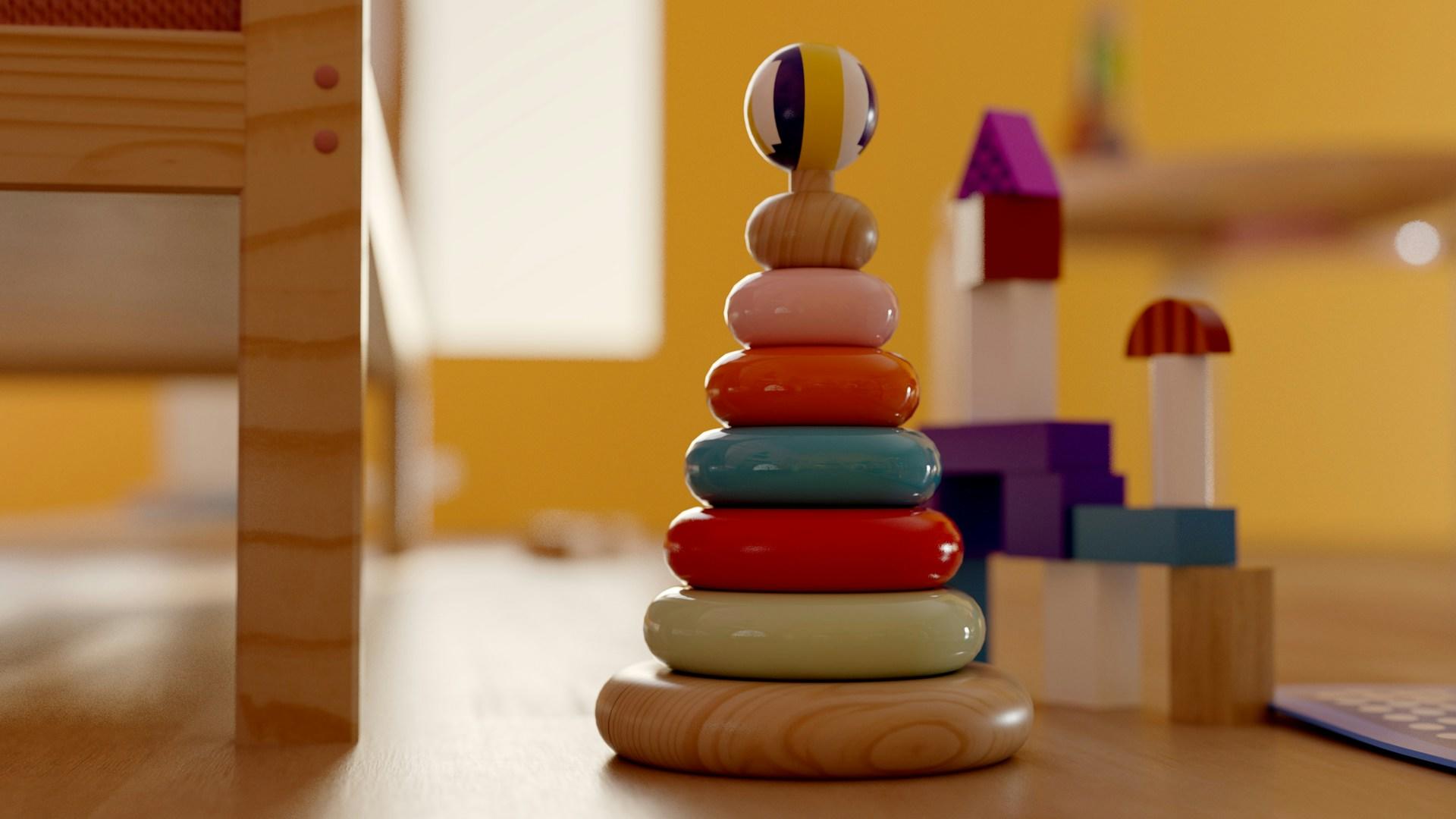 A set of colourful staking rings sits on the floor in front of a structure of building blocks, next to a child's wooden bed in a bright yellow room.