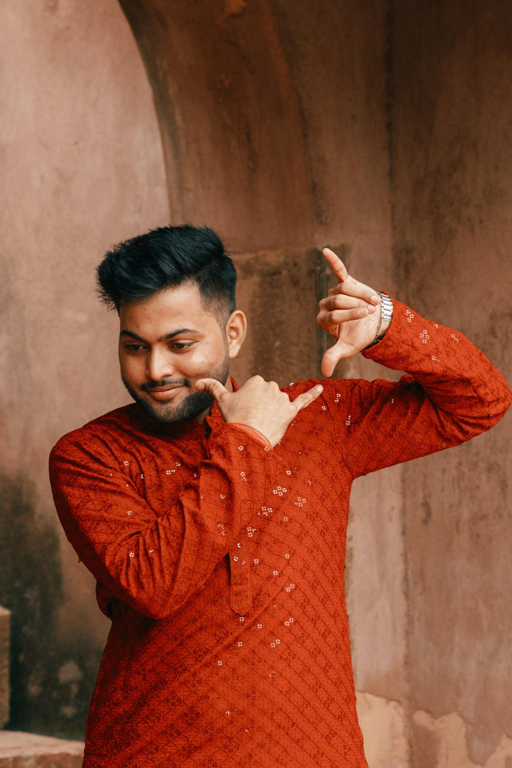 A person wearing a long-sleeved orange shirt with a shiny diamond pattern stands in front of a neutral brown background with their hands held at shoulder height, miming playing a wind instrument.