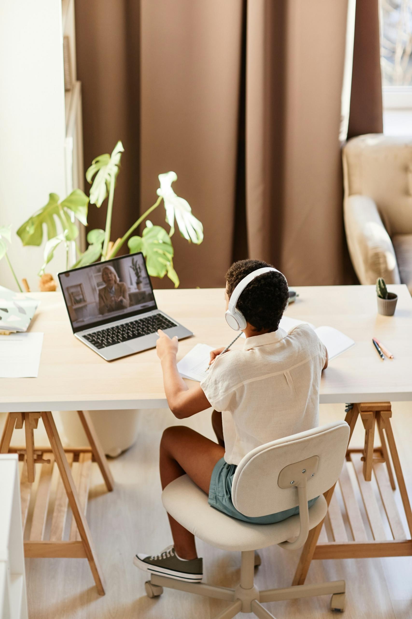 A person wearing a white top and denim shorts sits at a white trestle-top table in a white office chair with white headphones on. They hold a pen in their right hand, poised to write in the notebook in front of them. Their left hovers over near the open computer's keyboard while the screen displays a person wearing business attire.