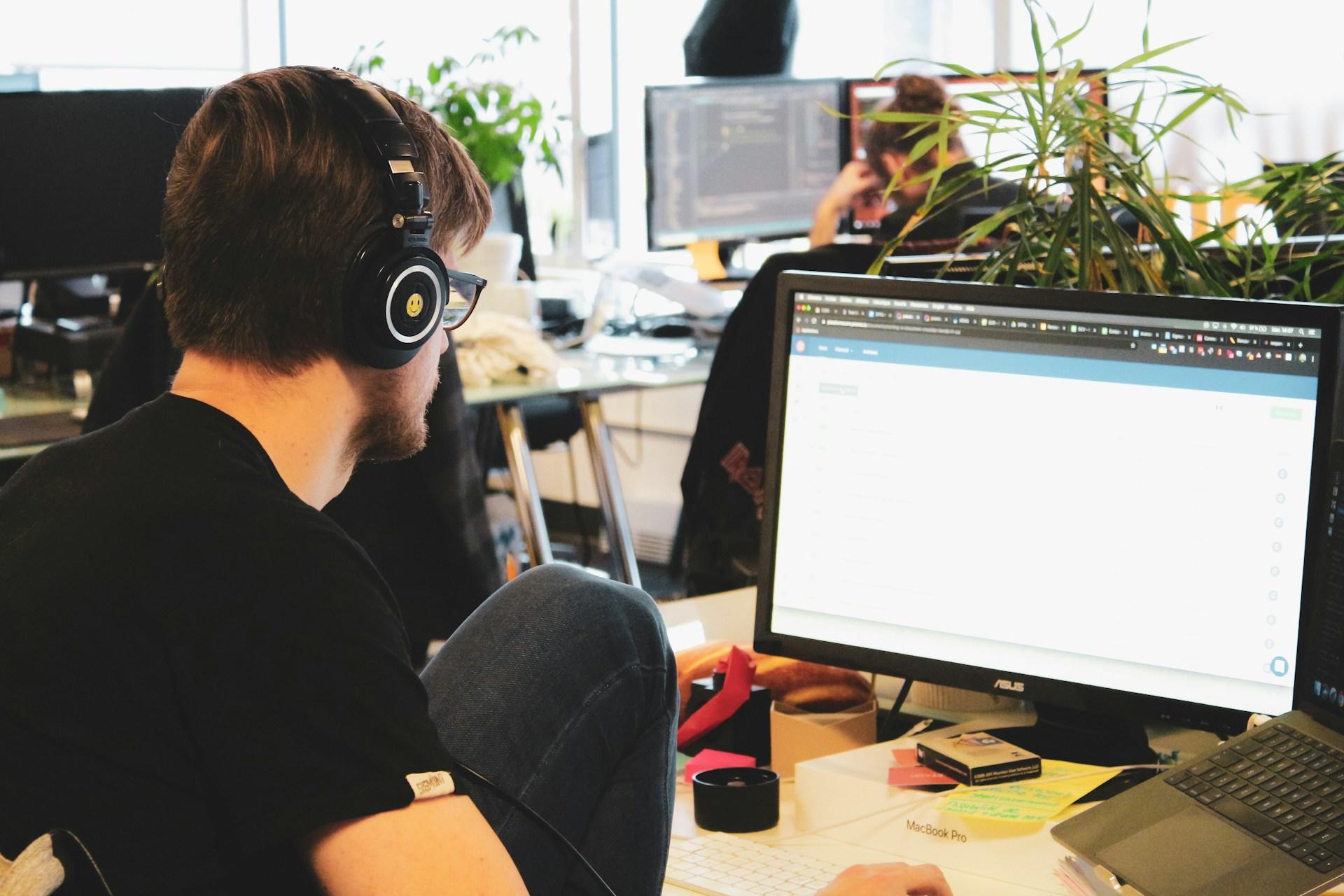 A person wearing a black tee shirt with black headphones on sits at an office workspace in front of a large computer monitor, which sits atop a messy desk.