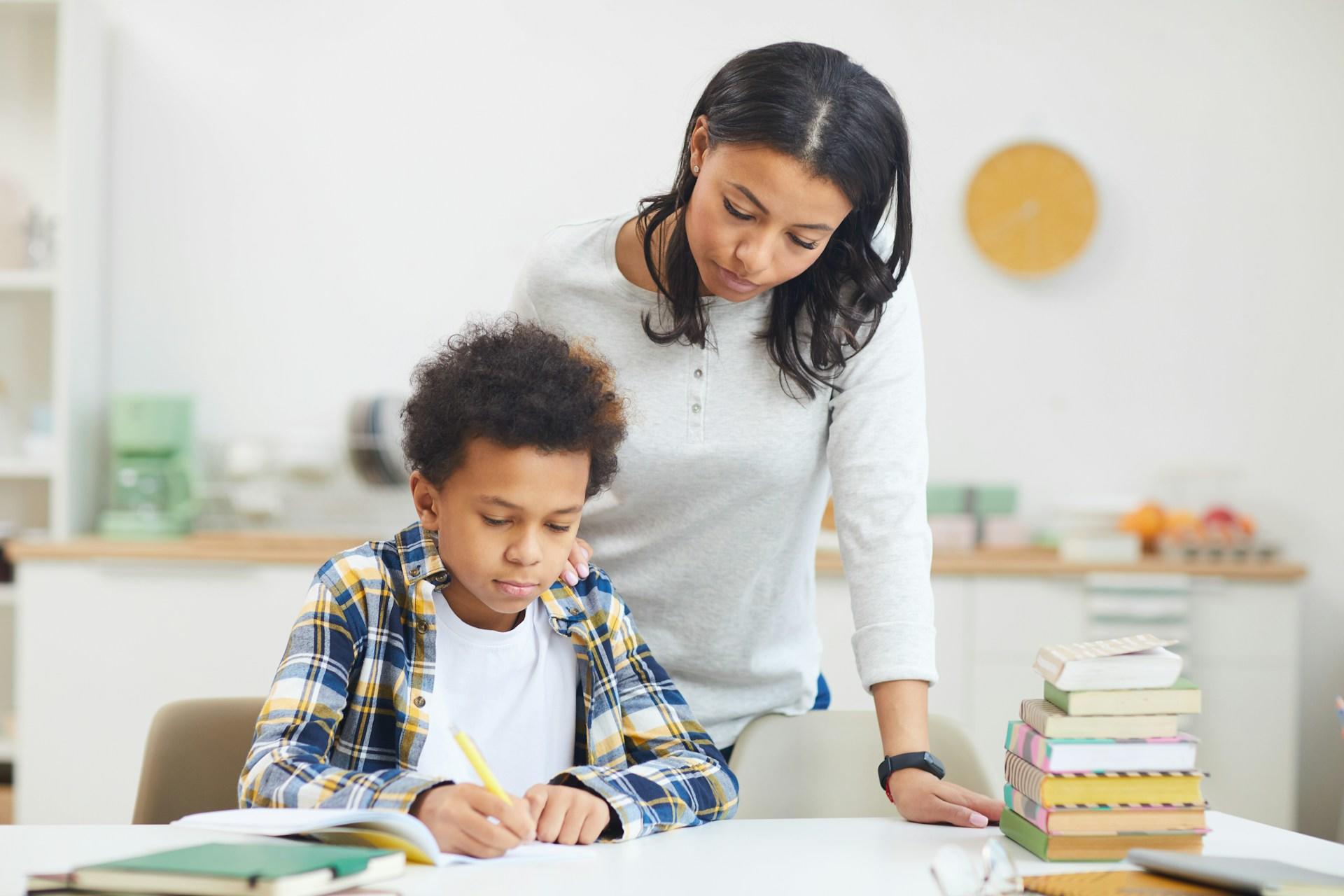 a child working on a desk with his mother hovering over