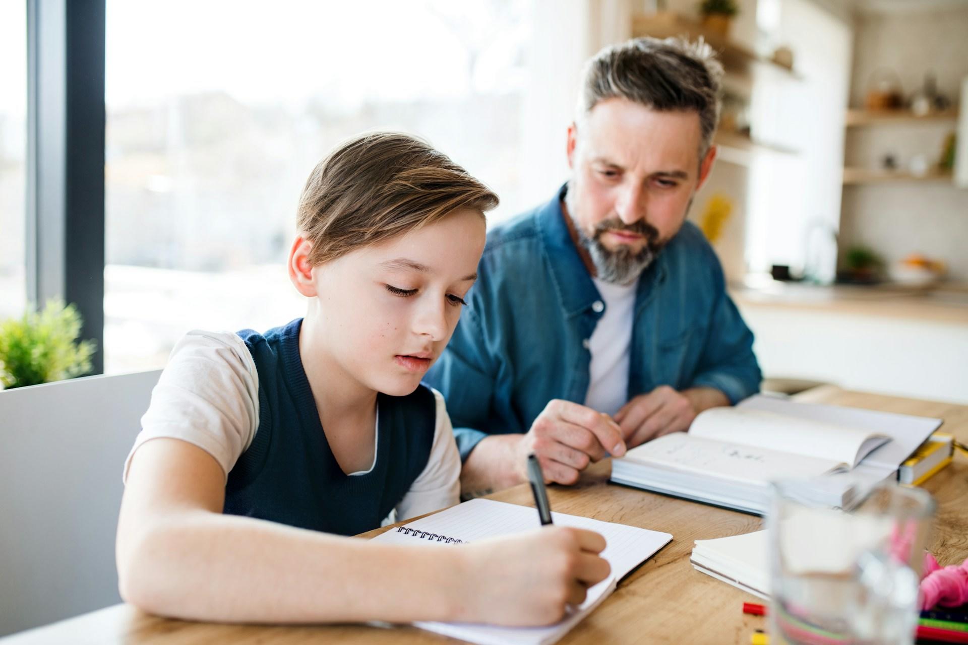 An adult wearing a teal shirt over a white tee sits at a table with a youth wearing a dark blue vest over a white tee, writing in a notebook as the adult looks on.