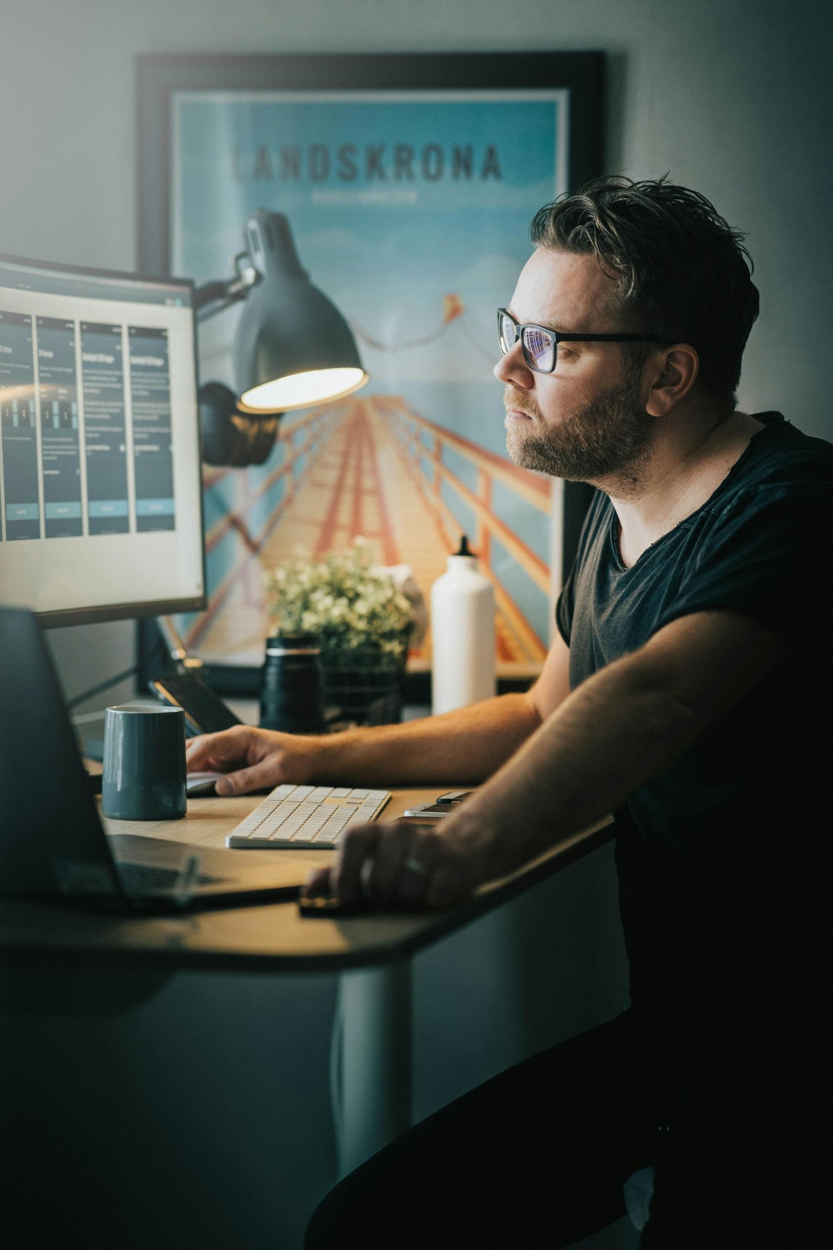A person wearing a dark coloured tee shirt and dark framed glasses sits in a darkened room at a desk with a large monitor on it and a desk lamp brightening the space.