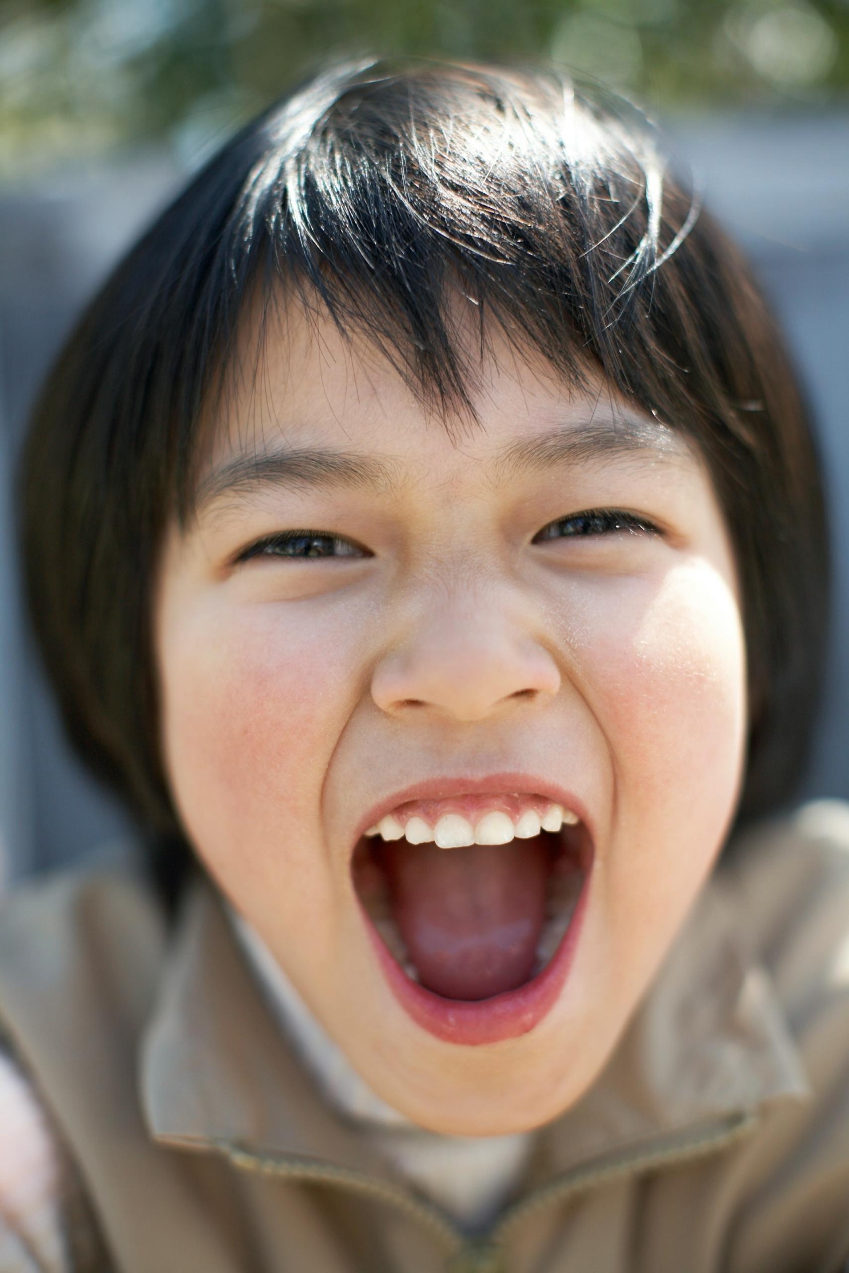 A child wearing a tan zip-up jacket over a white shirt stands outdoors on a sunny day with their mouth wide open, displaying white, even teeth.
