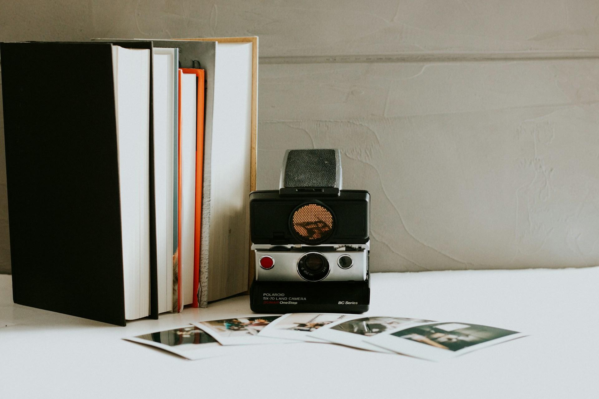 A vintage Polaroid camera next to stacked books, with several instant photos spread out on a tabletop.
