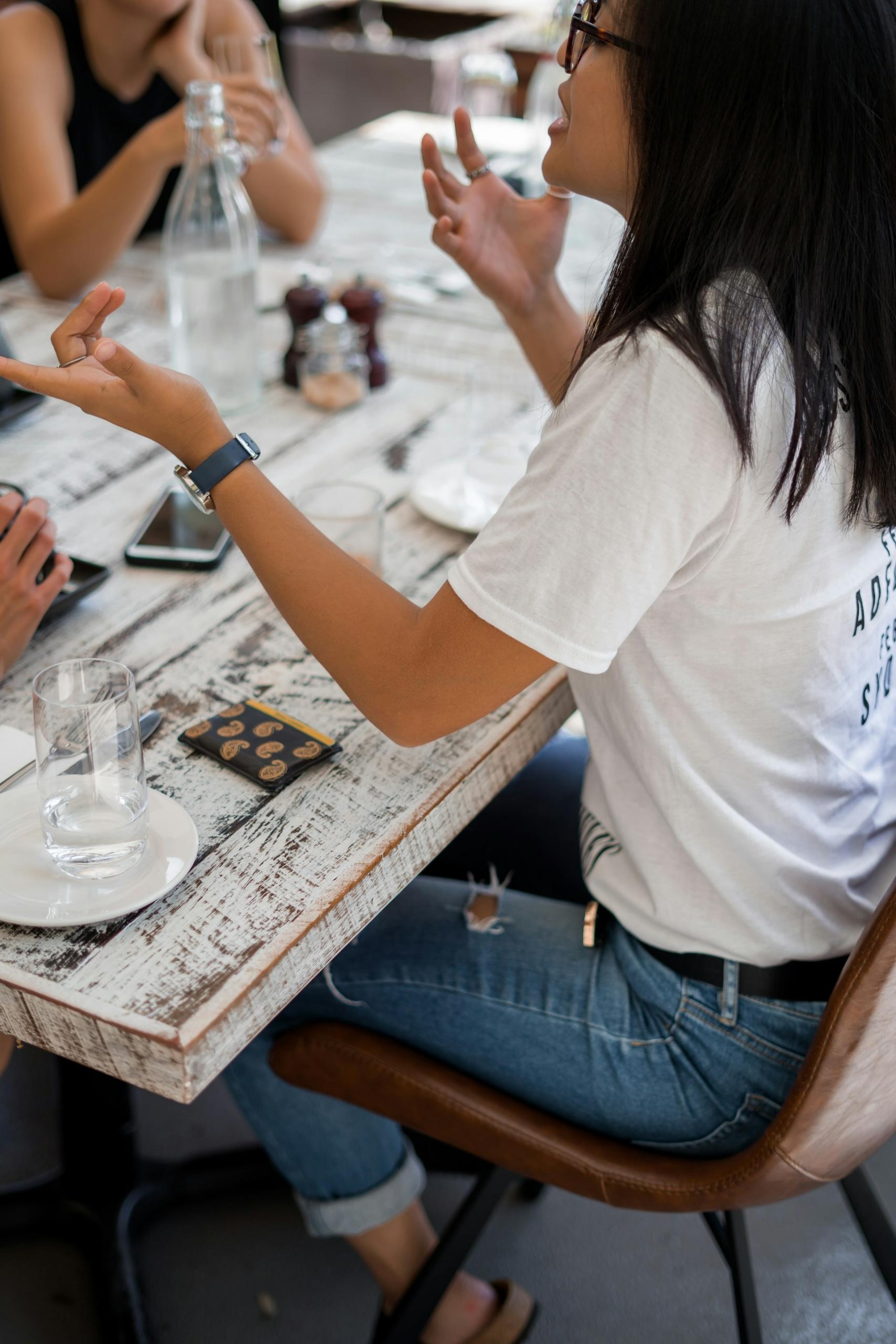 A person wearing a white tee shirt and blue jeans, with dark framed glasses on their face, sits at a table opposite at least two other people, holding their hands up as their talk. 