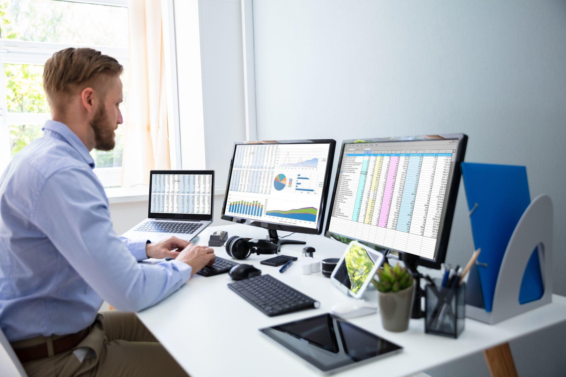 A man looking at three computer screens with spreadsheets on them
