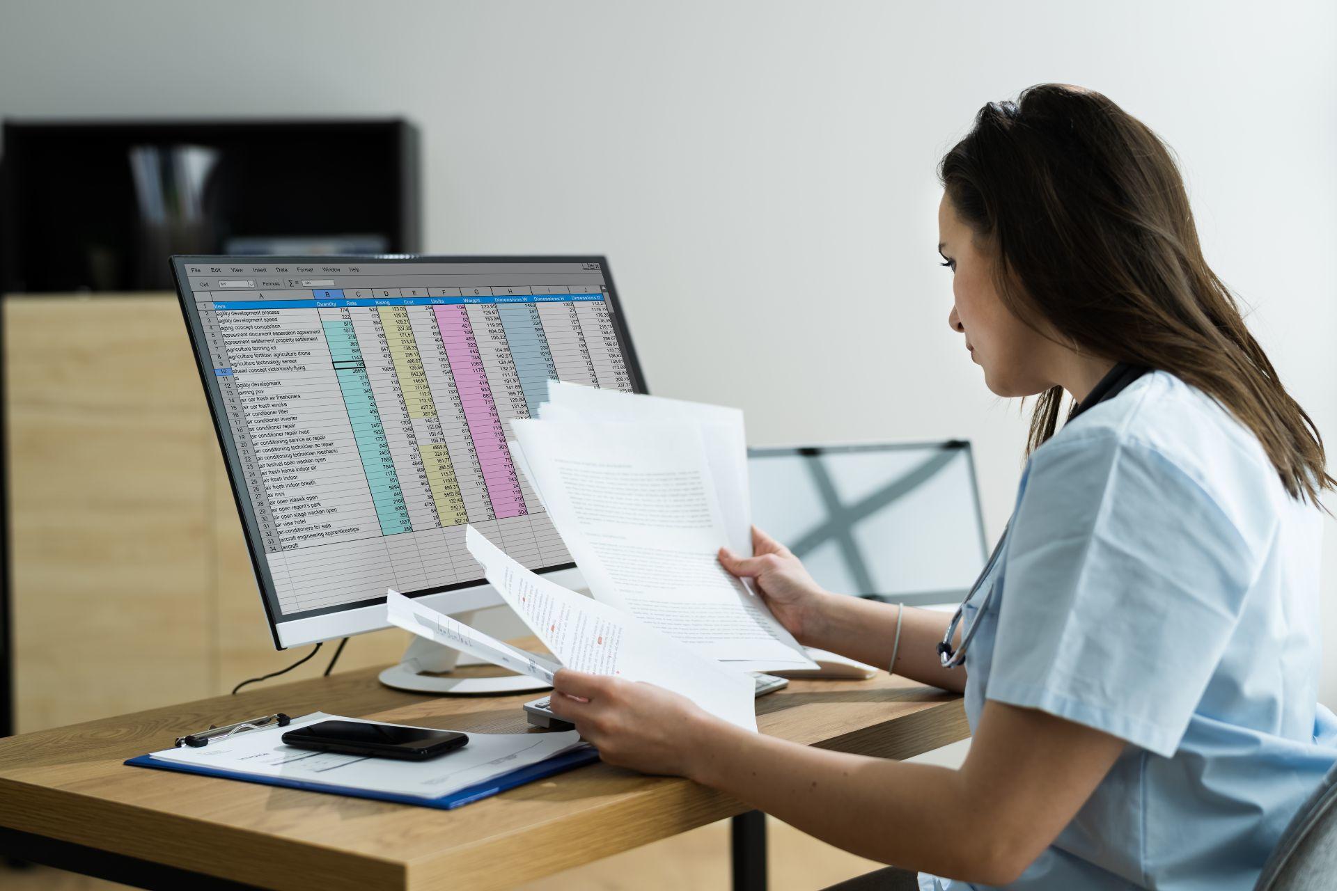 A person with Excel on their computer screen and looking at printed papers.