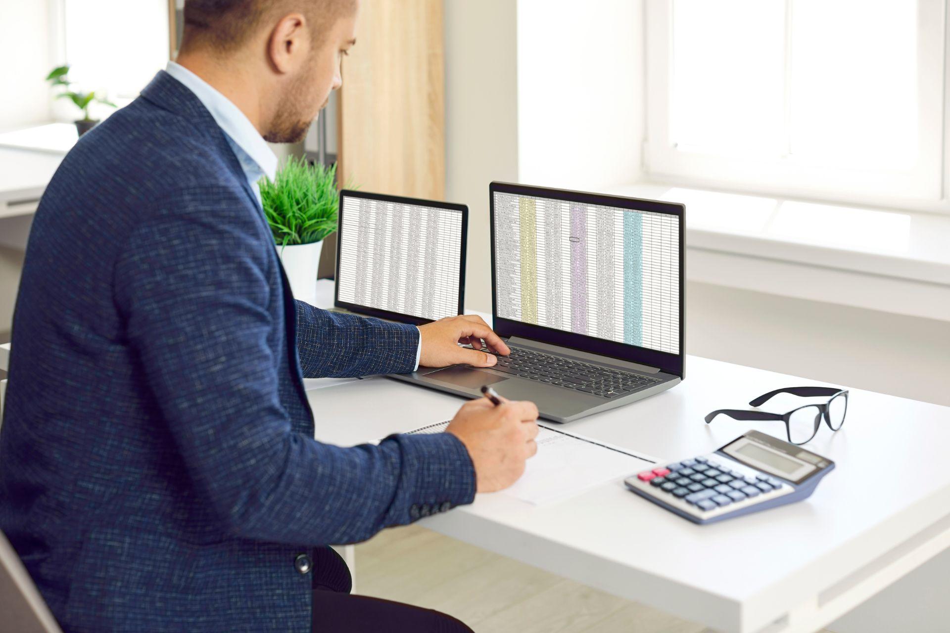 a man using Excel on two laptops at a desk with a calculator nearby