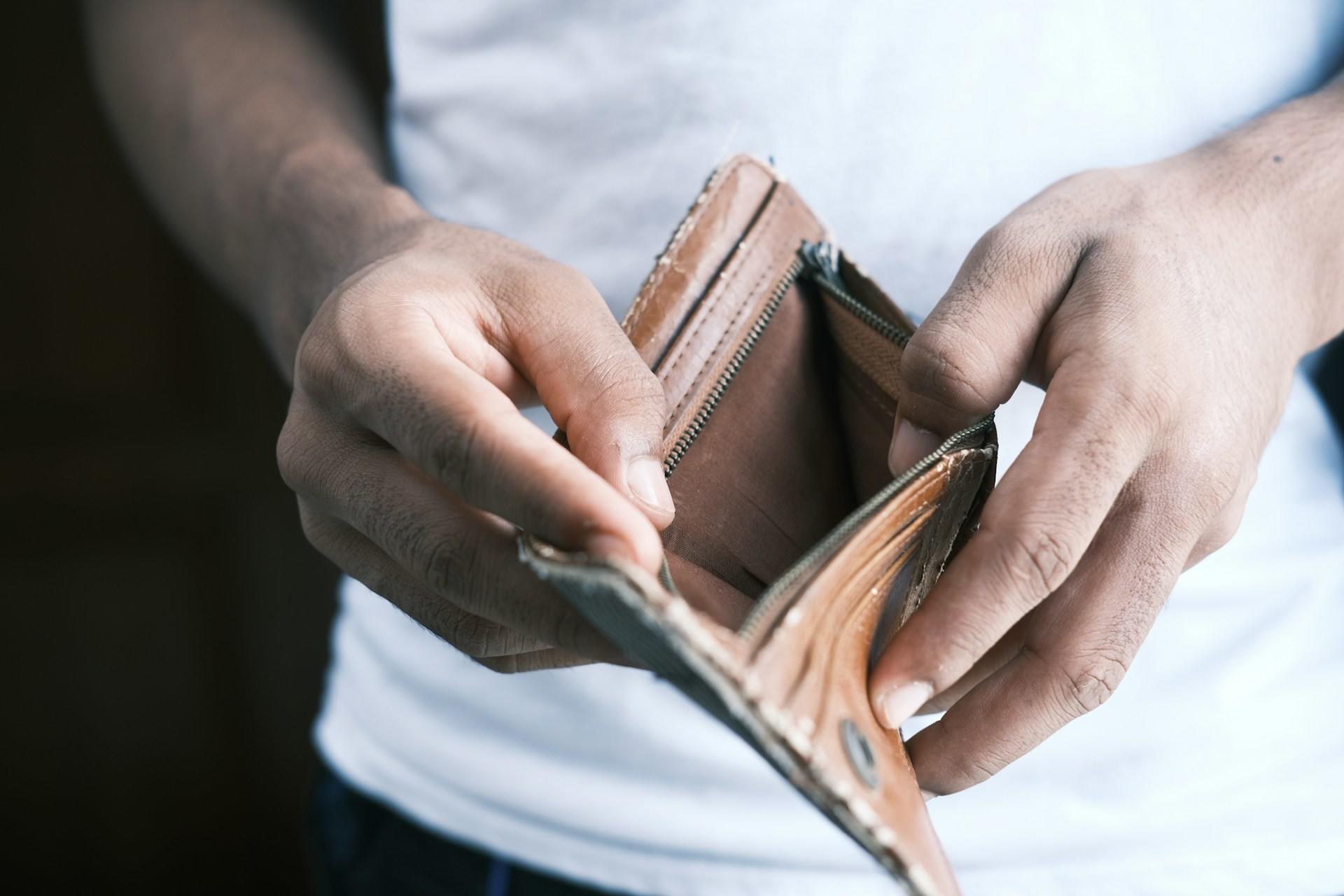 A person wearing a white tee shirt holds a brown wallet in both their hands, pulling it open to reveal it is empty.