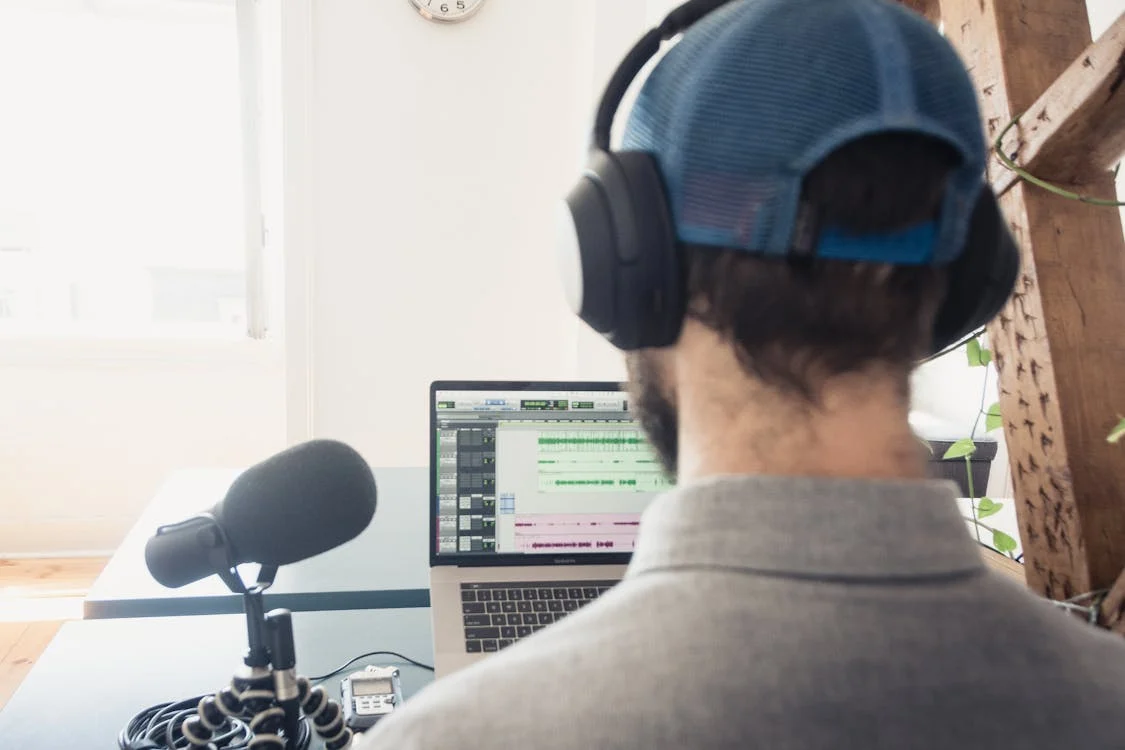 A person wearing a blue hat and a headphone in front of a laptop and microphone.