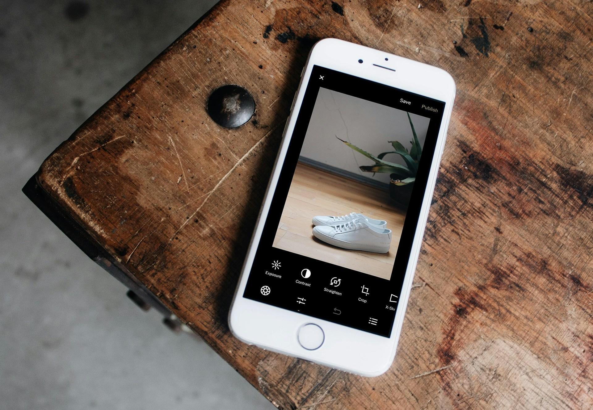 A smartphone displays a photo of white sneakers on a wooden table.