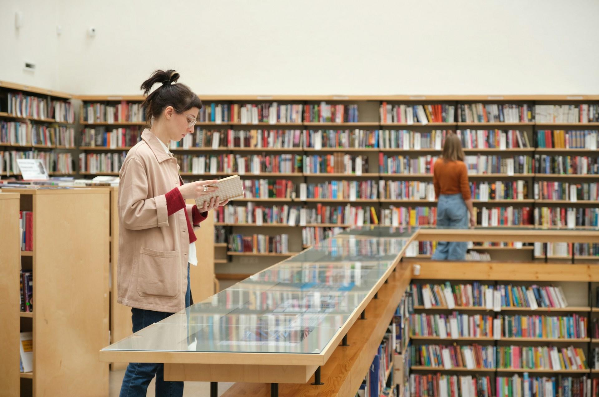 A person in a library, wearing glasses and a light-coloured jacket over a red shirt holds an open book, looking down on the pages while another person wearing a burnt orange top and light blue denims stands in front of the bookshelves, contemplating titles.