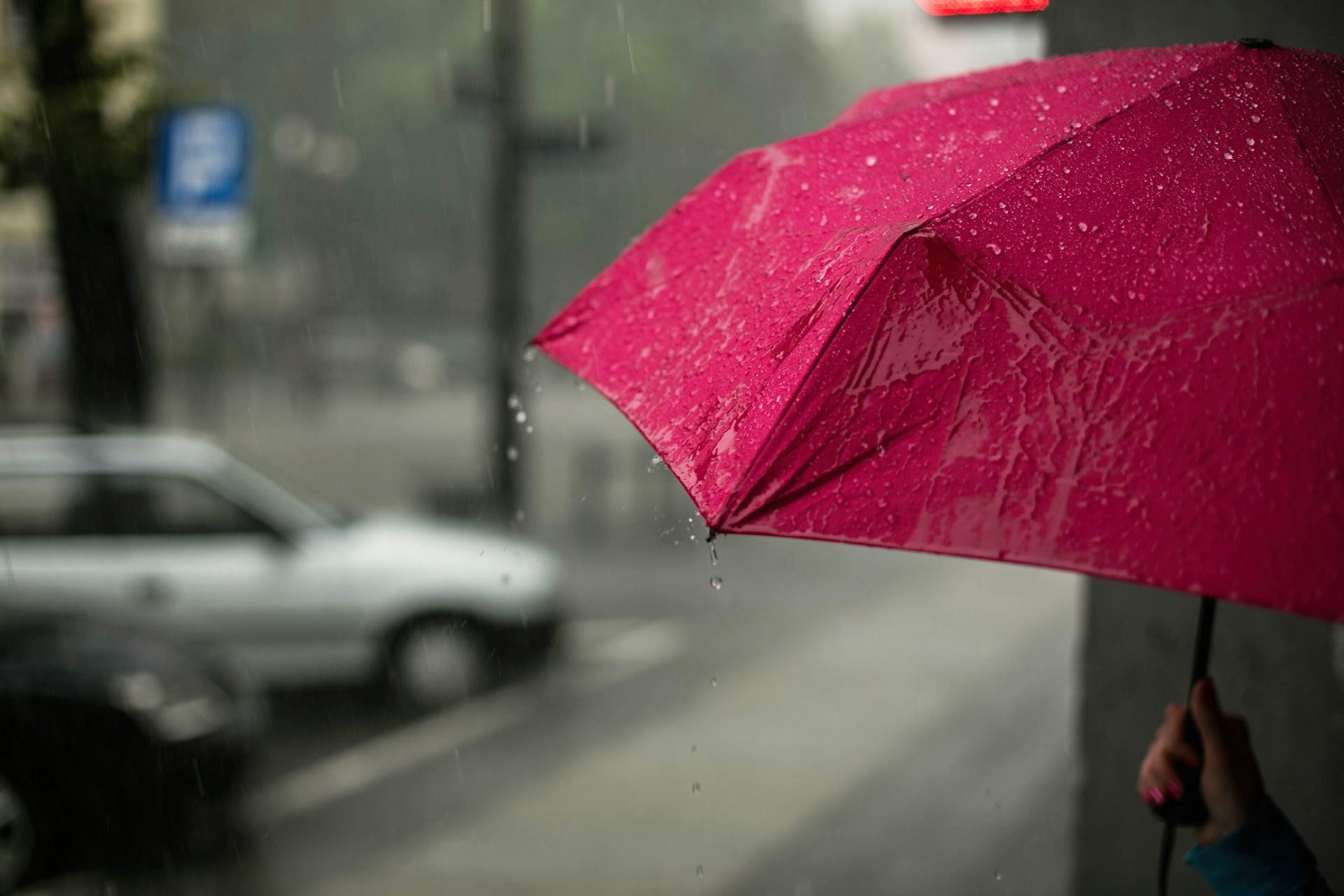 A person holding an open pink umbrella against the falling rain shelters in a building's alcove against a blurred backdrop of a grey city scene where you can just make out a blue and white parking sign.