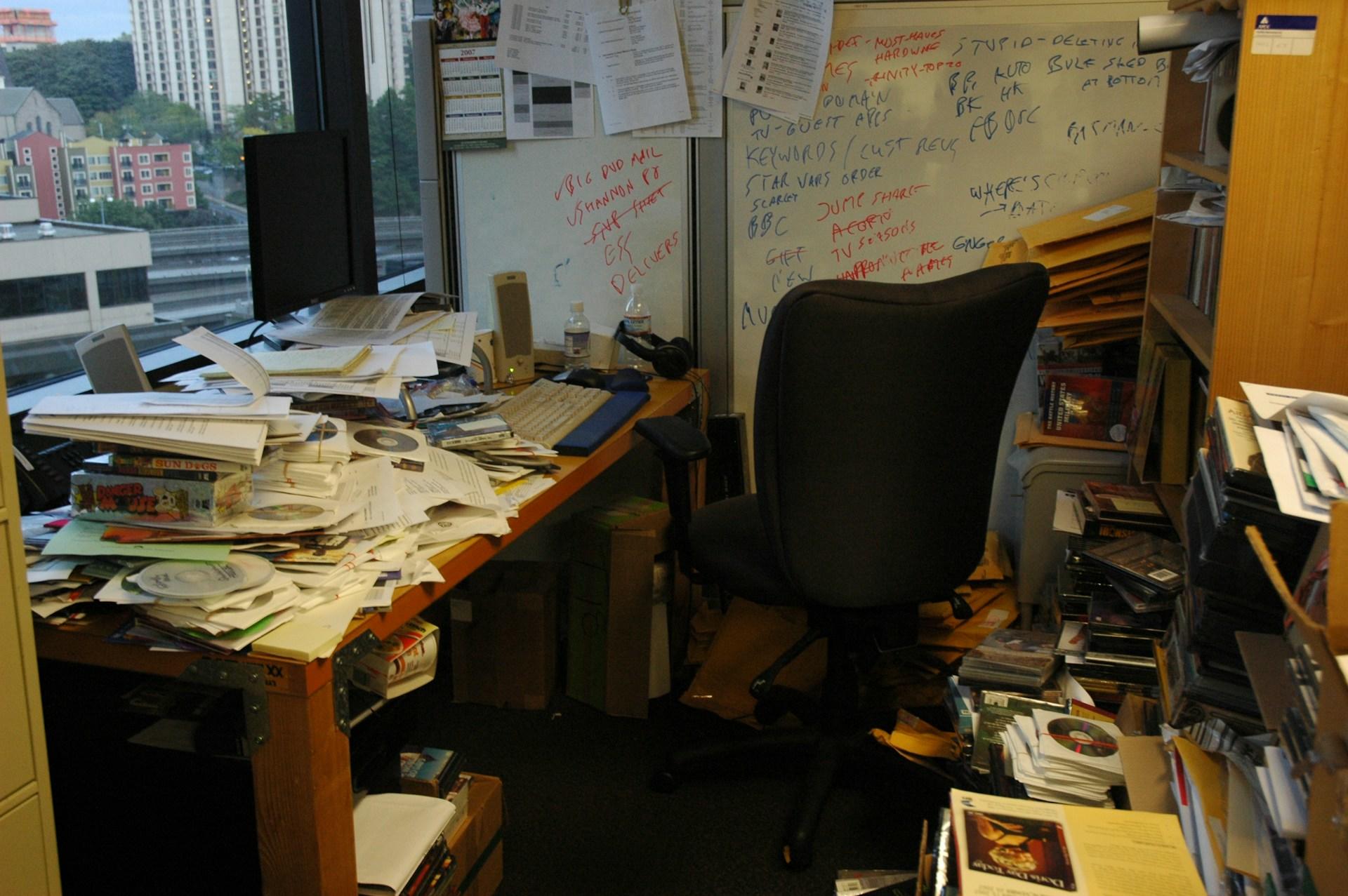A wooden table piled high with assorted papers, boxes and compact discs, with a computer sitting on the far end. A black office chair awaits its occupant as it sits in front of a dry-erase board with red and blue writings on it.