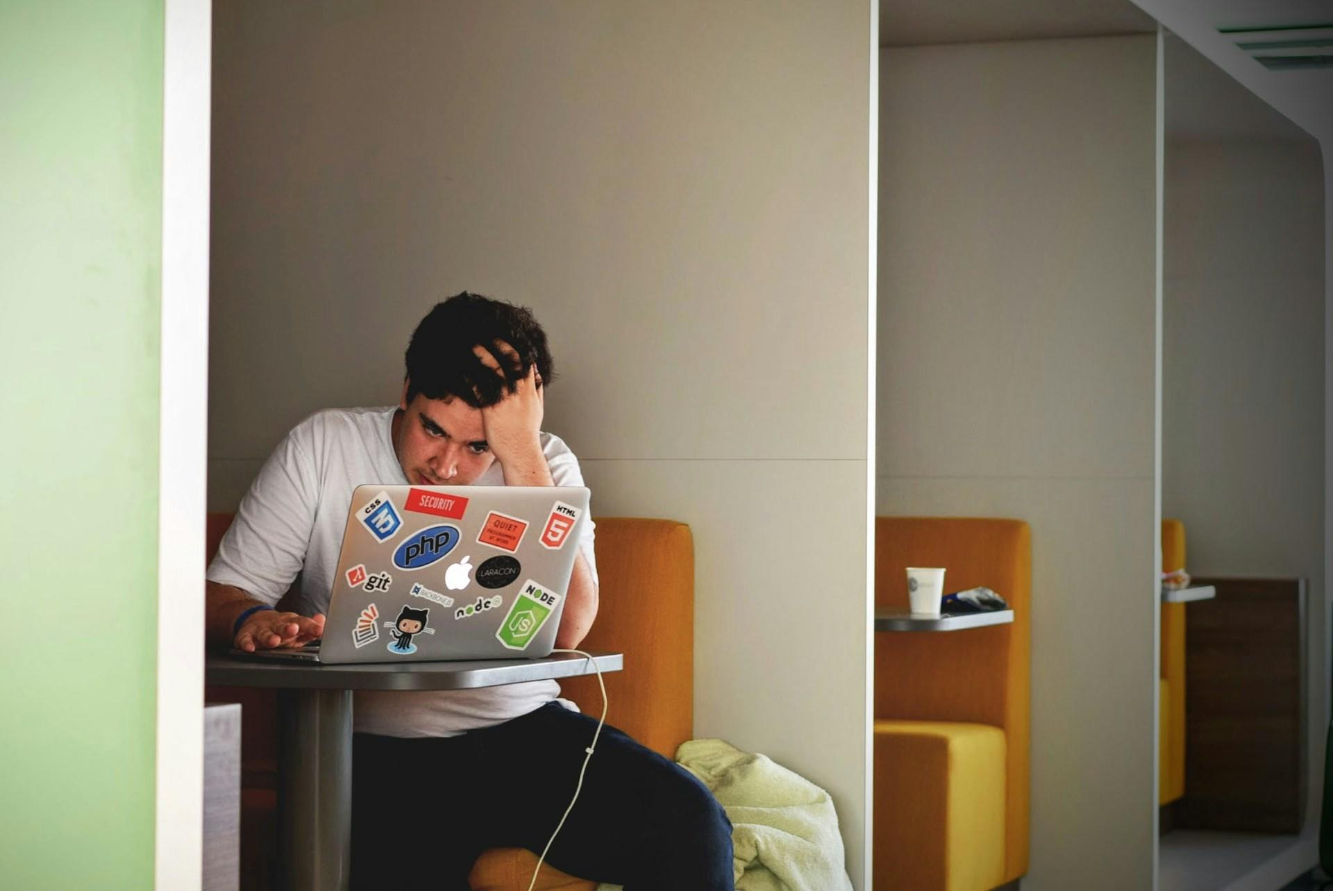 A person wearing a light coloured tee shirt and dark trousers sits in a booth on a light brown bench in front of their open laptop computer with their head propped on their left hand with a look of disbelief an anxiety on their face.