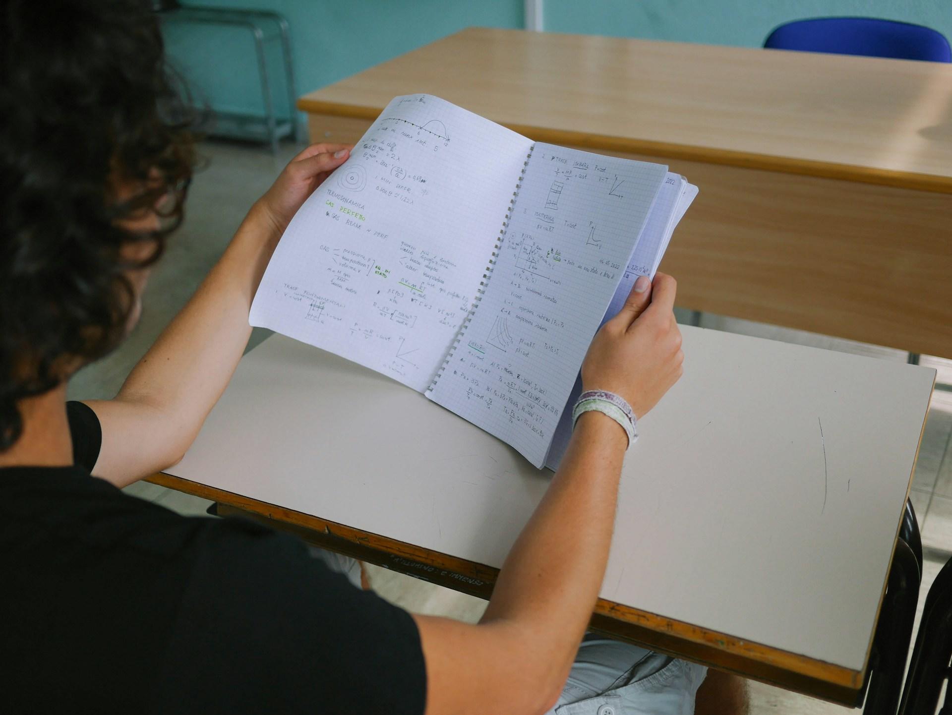 A person with curly hair, wearing a black short sleeved top sits at a school desk holding a spiral-bound notebook open in both hands, revealing both pages with writing on them.  