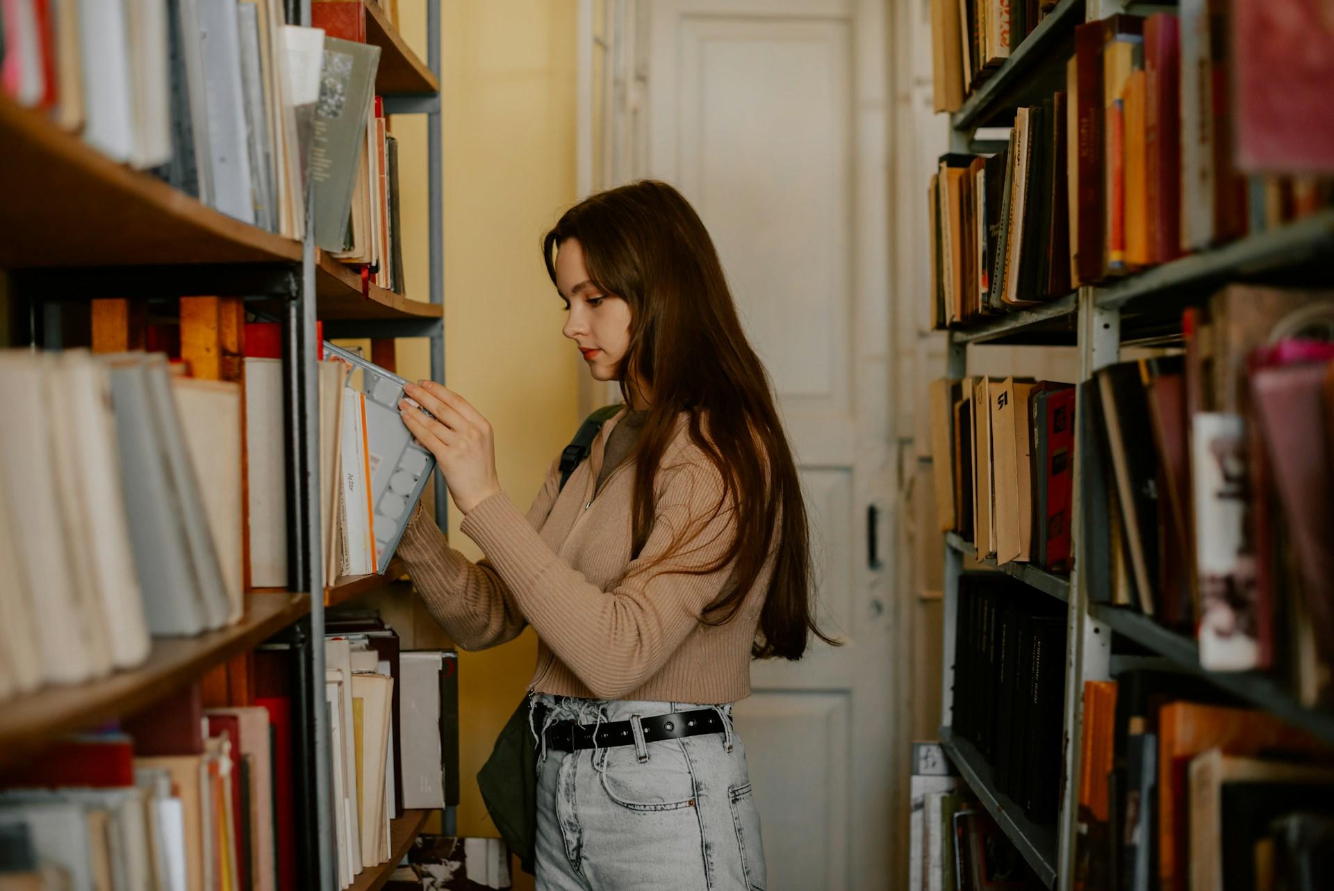 A person in a tan jumper and faded jeans with a black belt selects a book with a grey cover from the library stacks