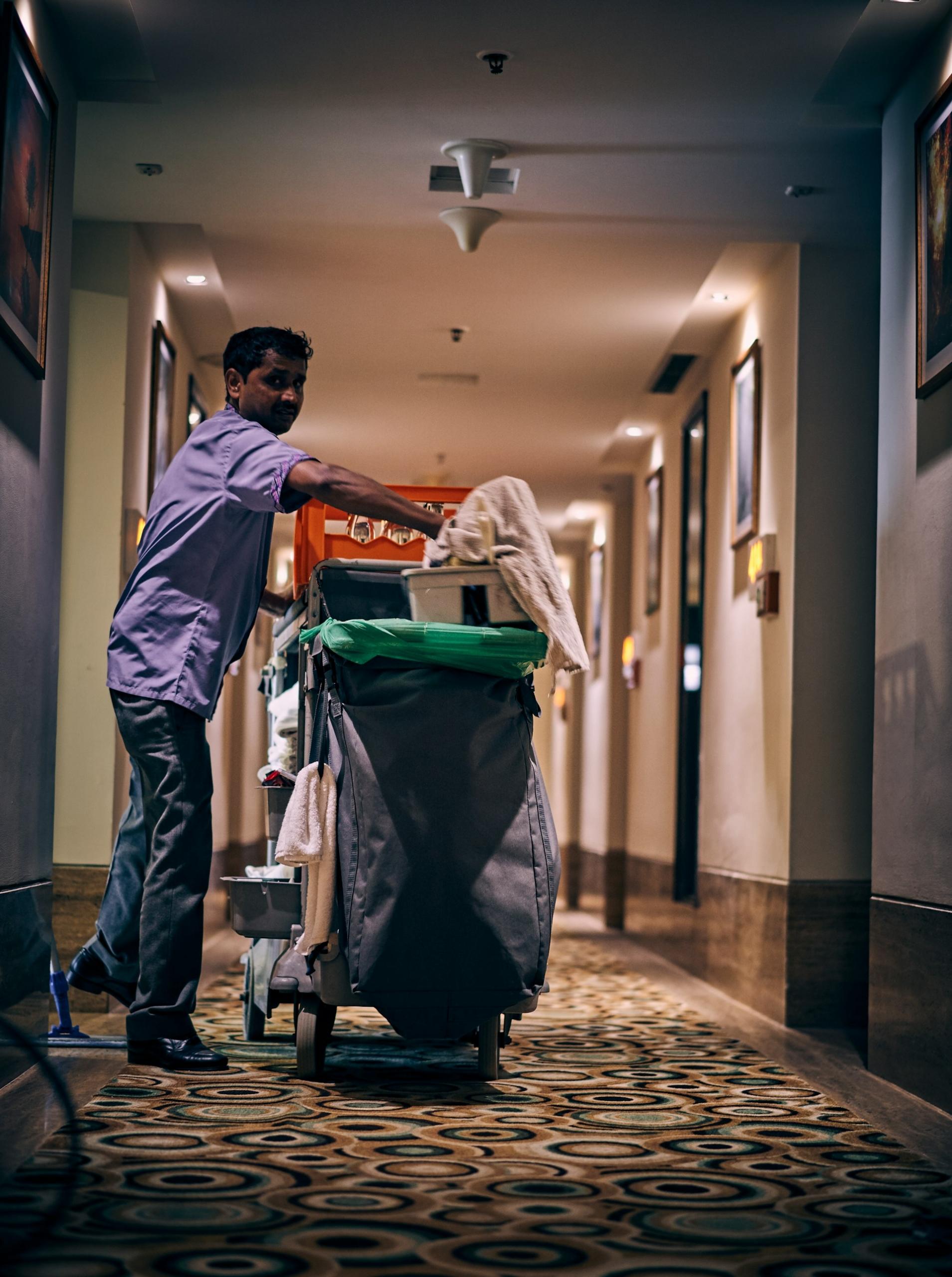A person wearing a blue shirt and jeans stands in a hotel hallway, next to a room service cart, reaching into the top, where a white towel rests, bunched up, on a white, compartmented container. 