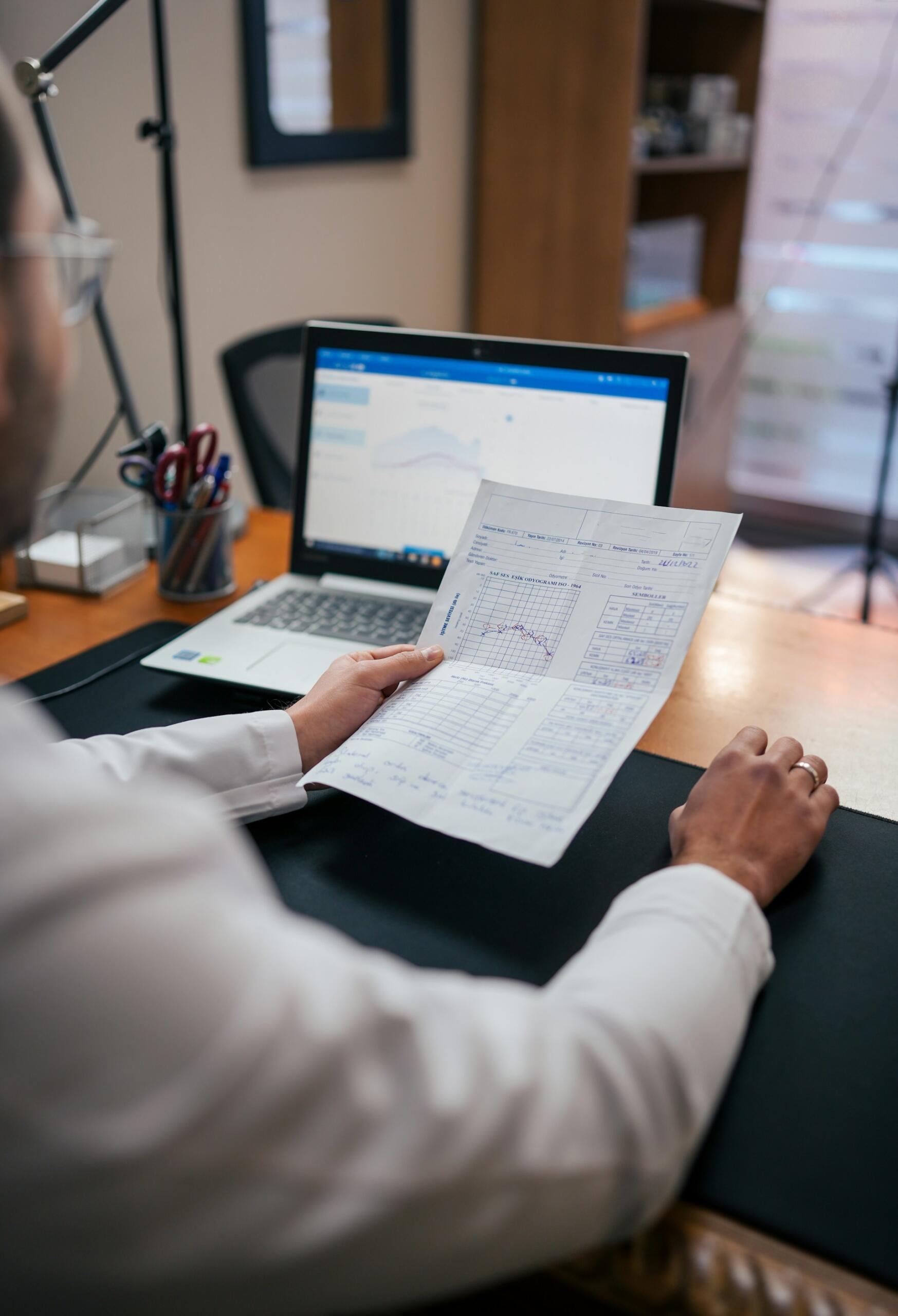 A person at a desk, with a paper displaying charts in front of a computer.