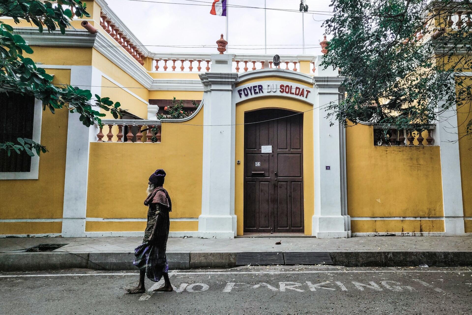 Person walking past a yellow and white colonial-style building on a tree-lined street in Puducherry