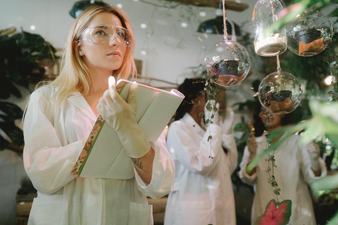 Female botanist in a lab coat studies plants and takes notes in a notebook.