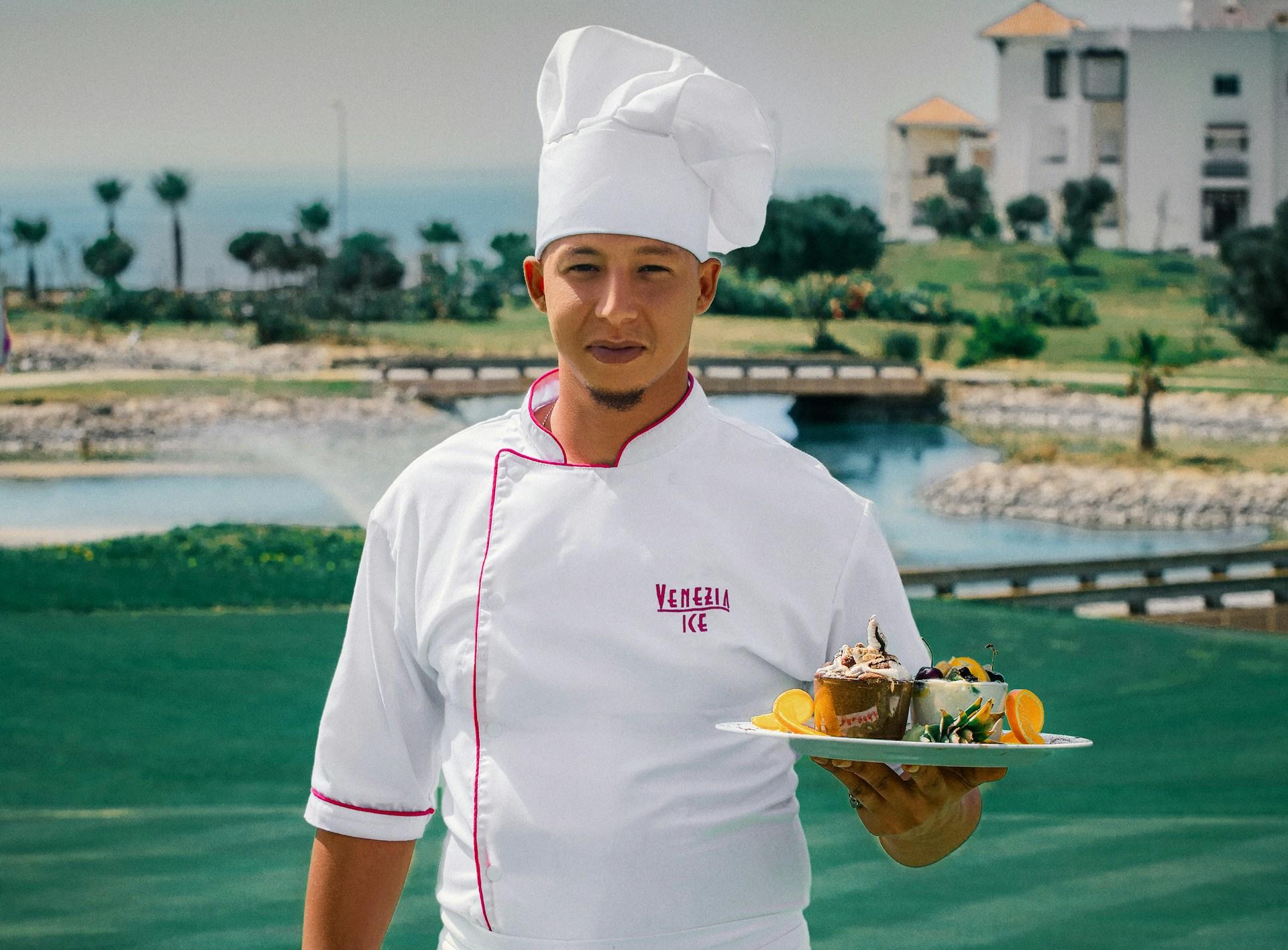 a chef in a white coat holding a plate with multiple deserts on it