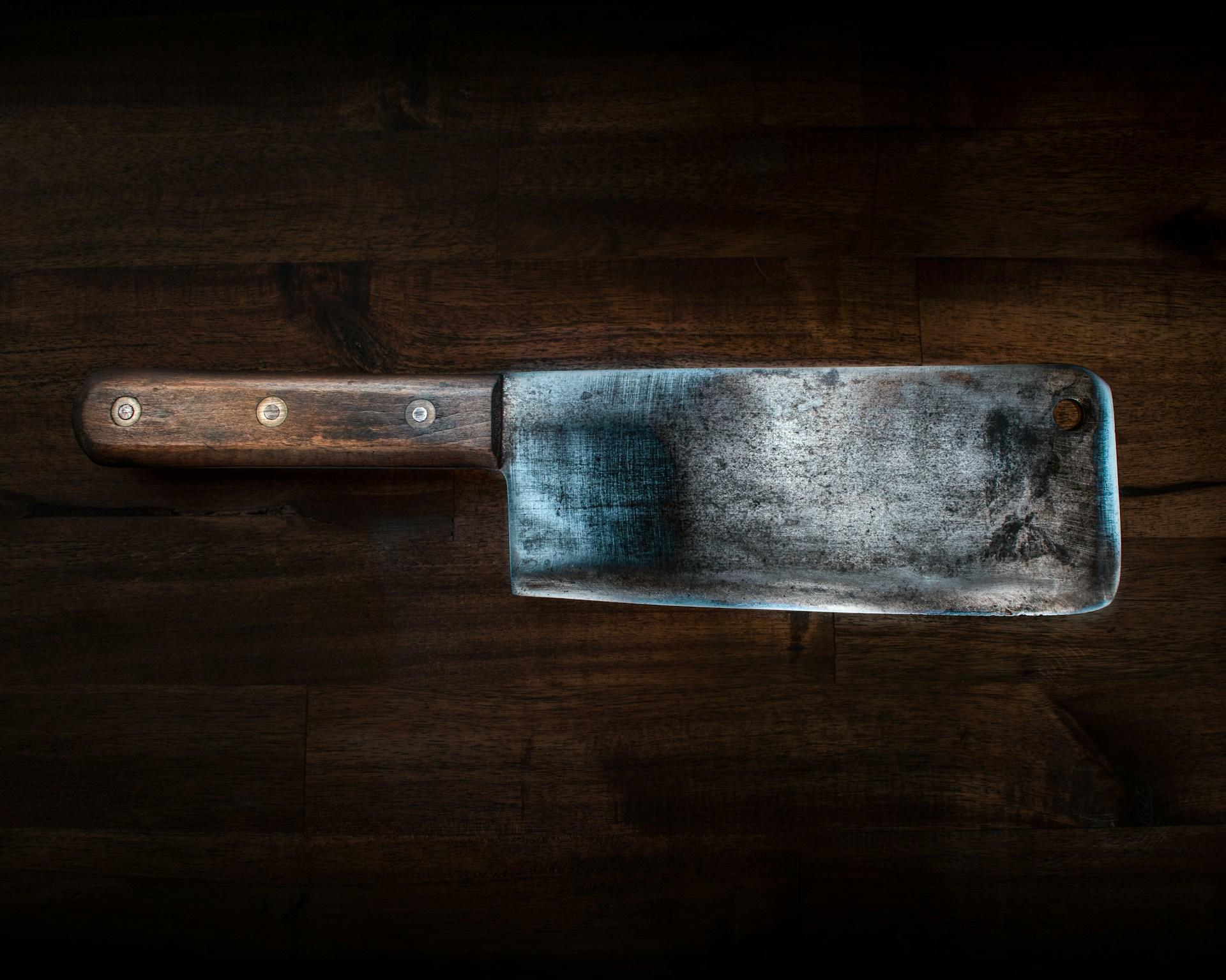 A big meat knife laying down on a wooden surface, waiting to be picked to work.