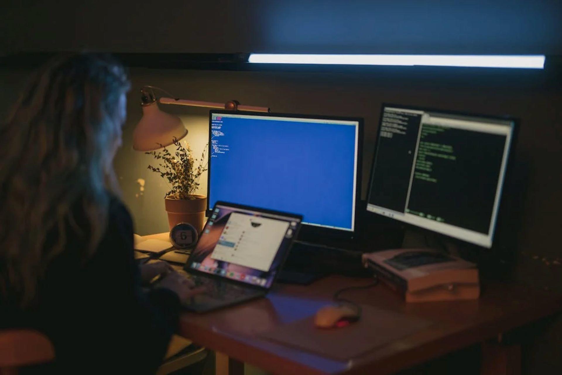 A person in a dim room sitting at a desk using many computers for coding.