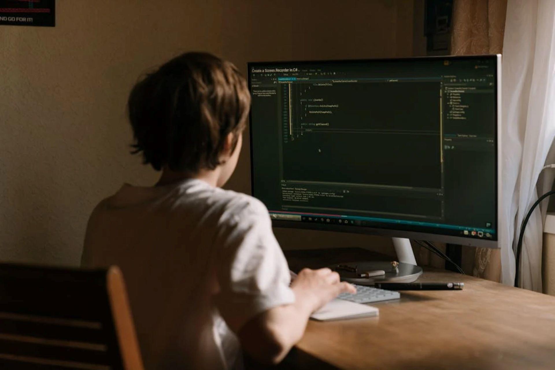 A young boy sits at a desk, focused on a computer screen displaying coding content.