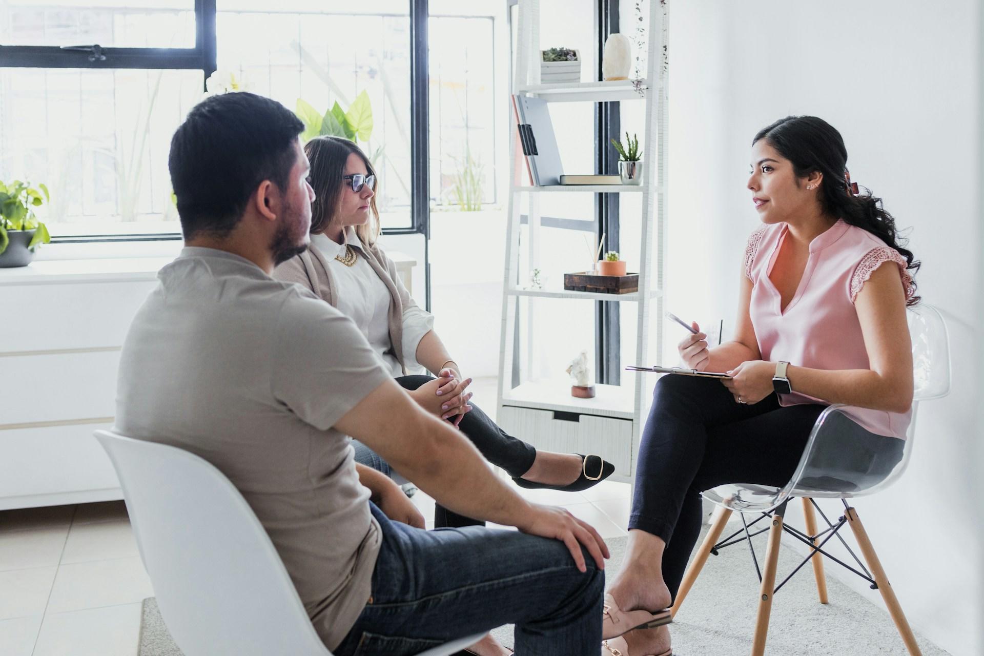 A person wearing a light coloured top and dark slacks holds a pen in their hand and a clipboard on their knee. they sit in a clear plastic chair, opposite two other people wearing roughly the same colours.