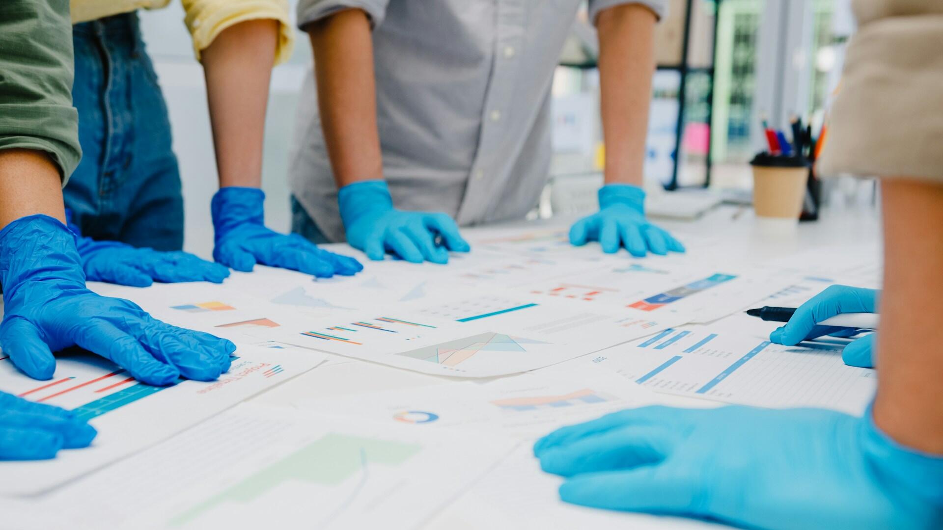 People wearing blue nitrile gloves lean on a table. 
