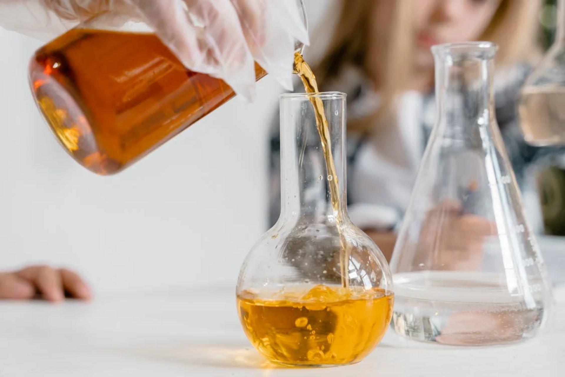 A person in a lab coat carefully pouring a yellow liquid into a beaker.