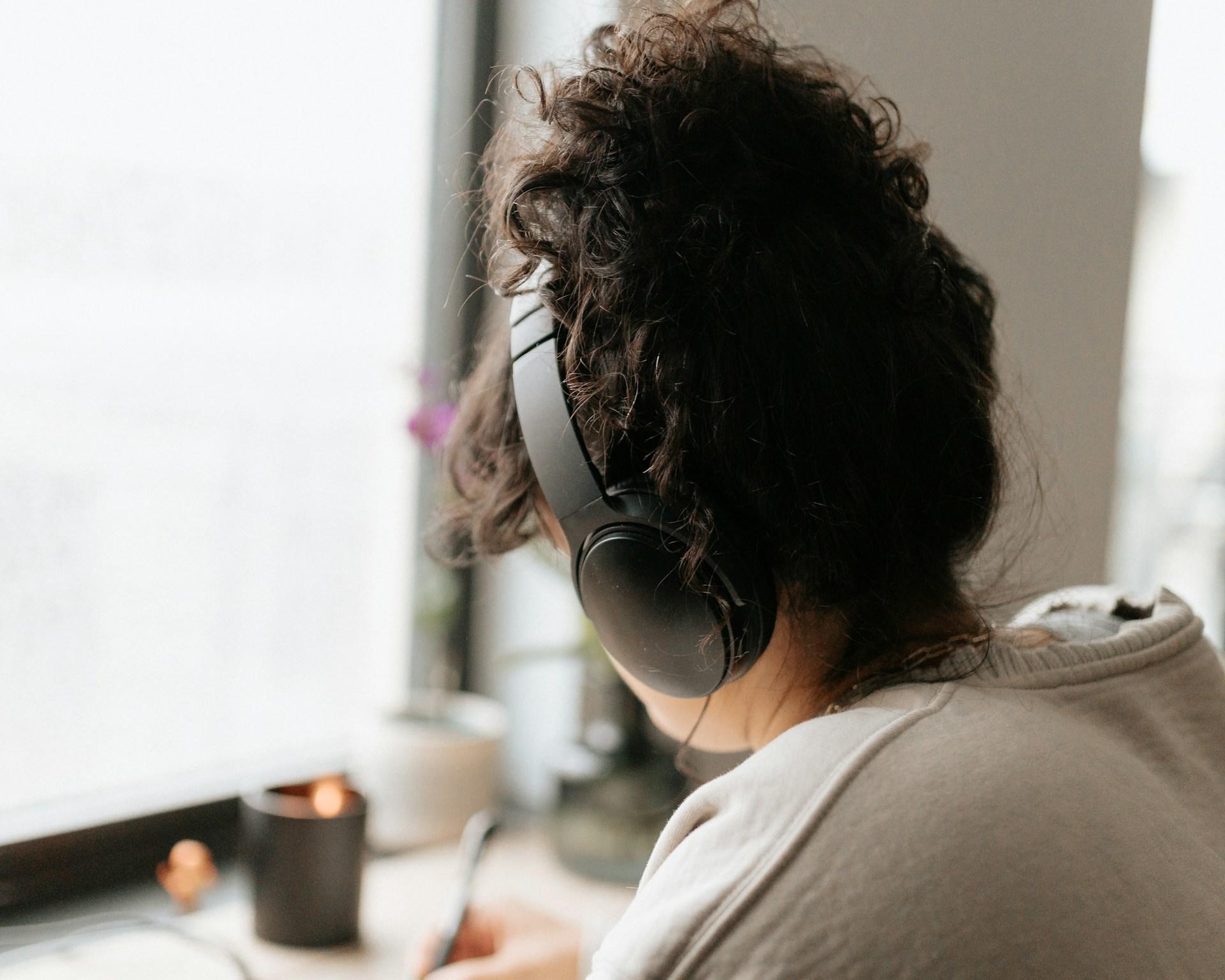 A person wearing a tan jumper and black headphones sits at a desk with a pen in their right hand. 