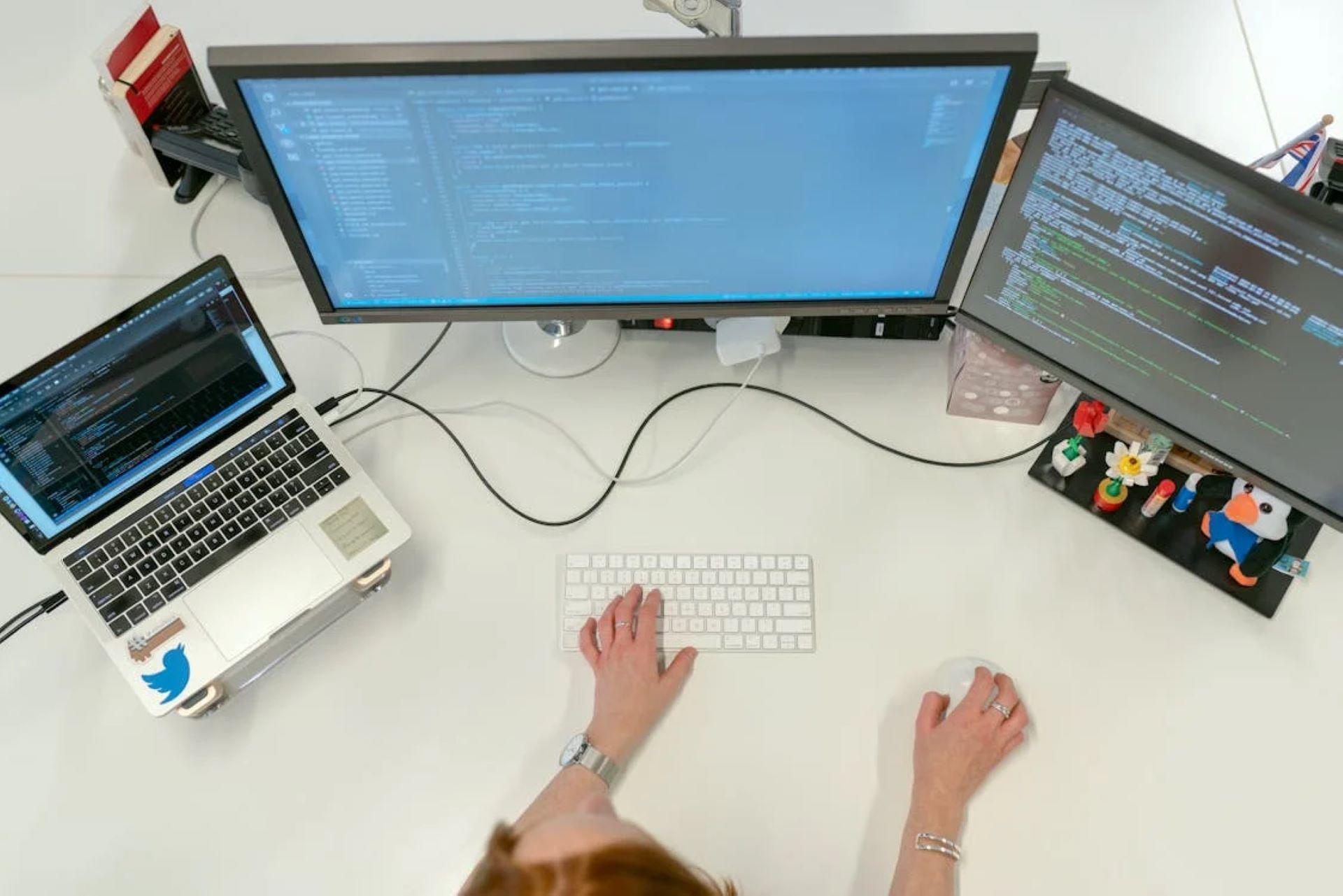 A programmer typing code on a keyboard with three computer monitors displaying code and data.