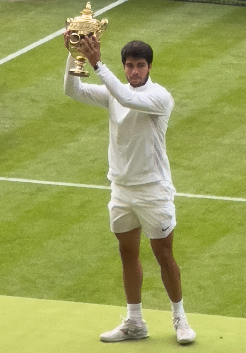 A person wearing white stands on a grass field holding a golden trophy cup aloft. 