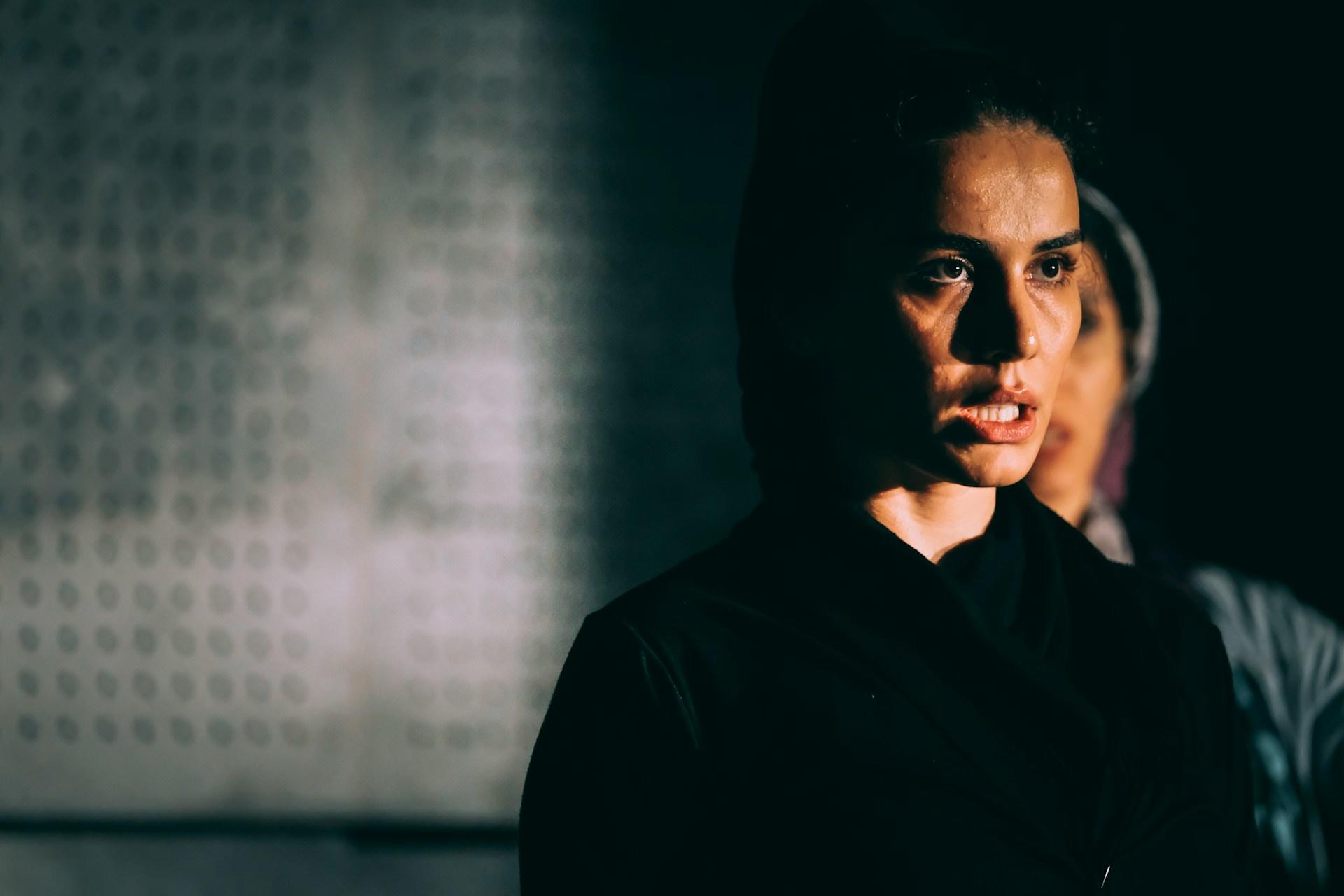 A person wearing black stands in a gloomy space with their mouth slightly open.