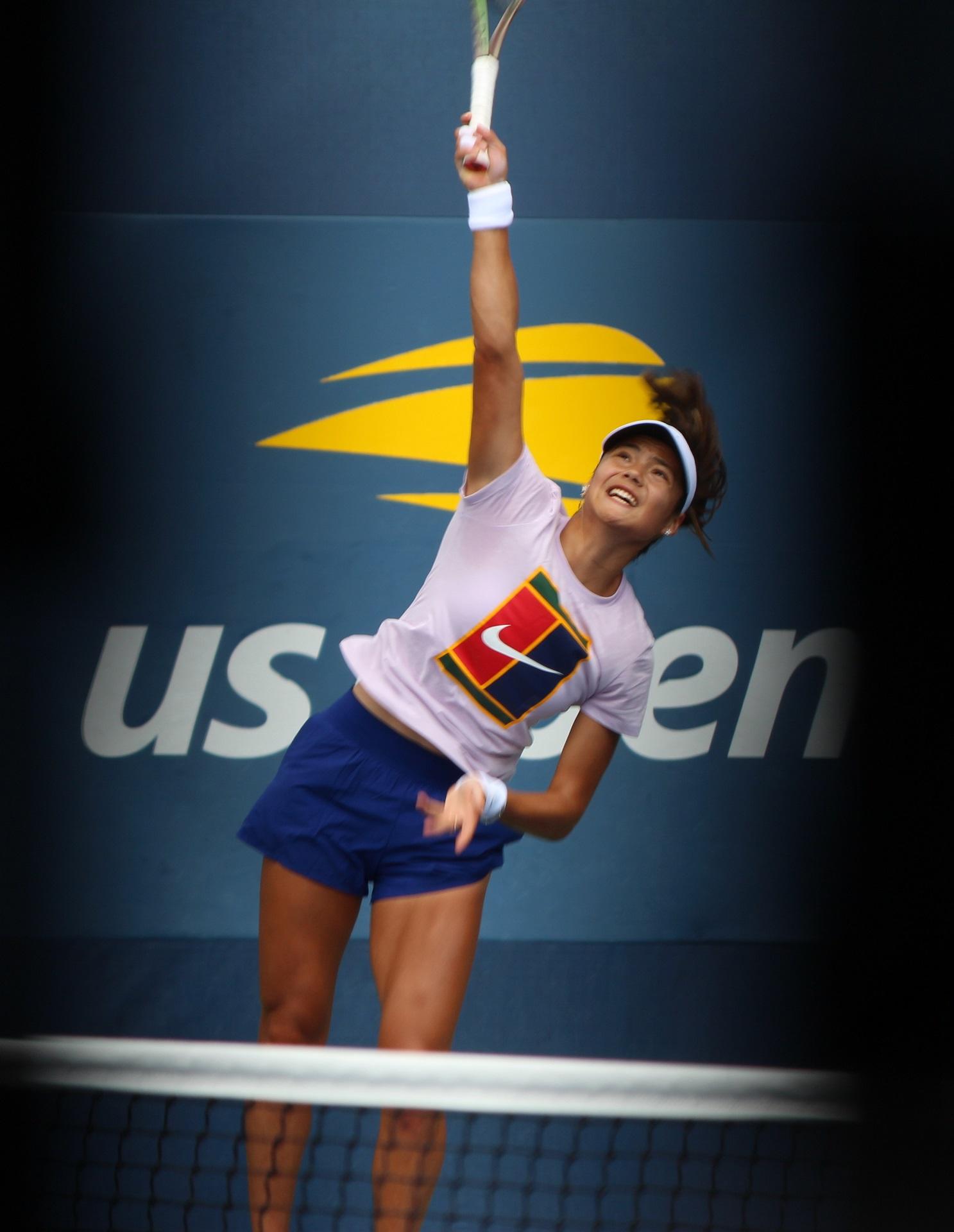 A woman wearing a white shirt with a logo and dark coloured shorts holds a tennis racket in the air. 