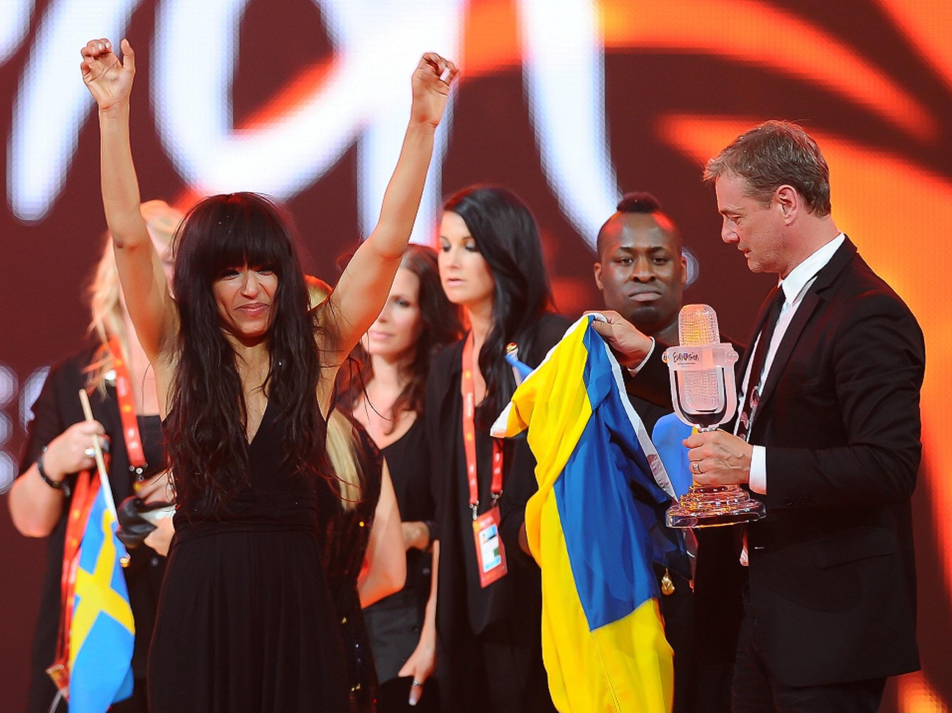 A woman wearing a black dress fronts a group of people against an orange and white backdrop.