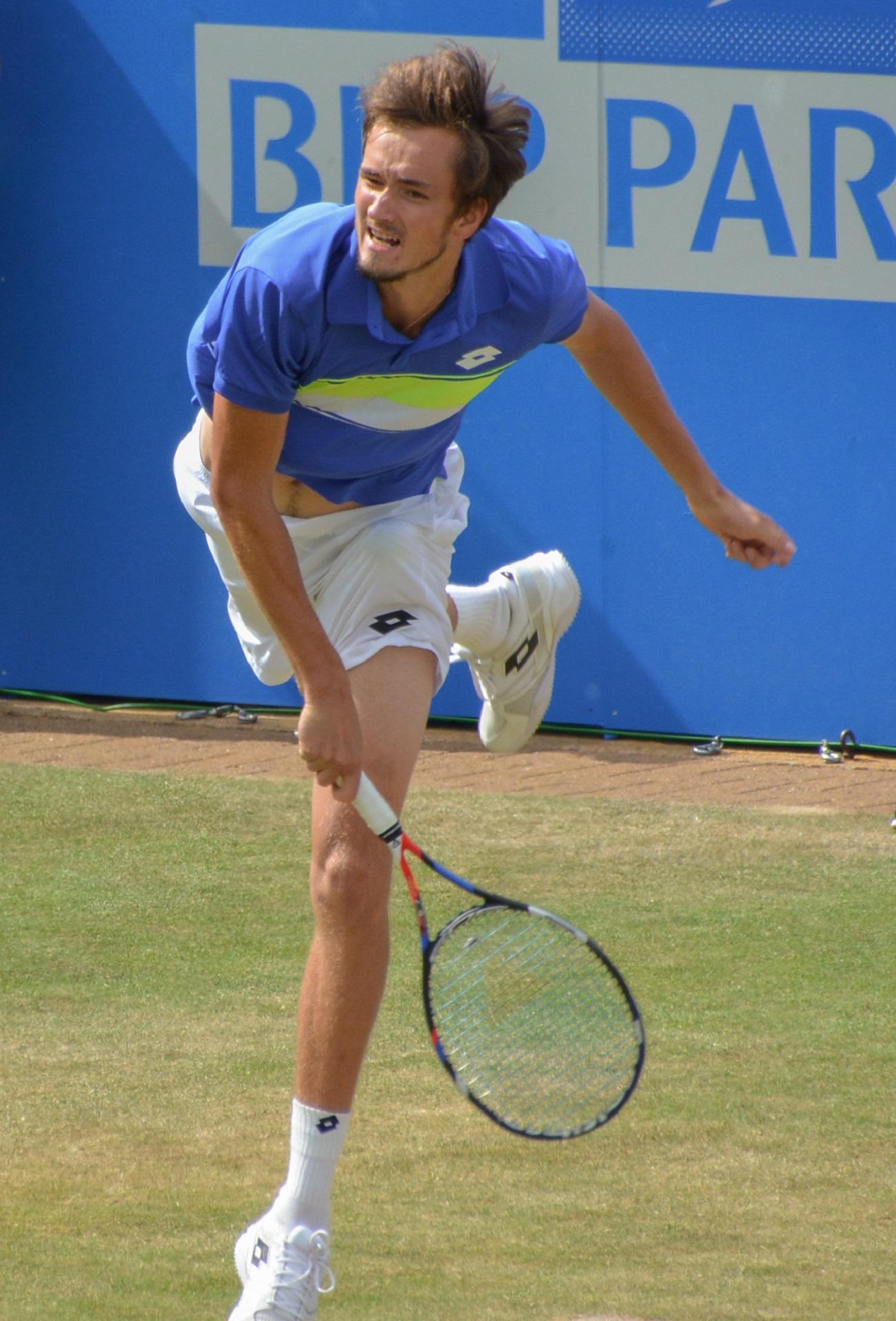 A person on a grass tennis court wearing a blue top with white shorts, holding a racket in their right hand. 