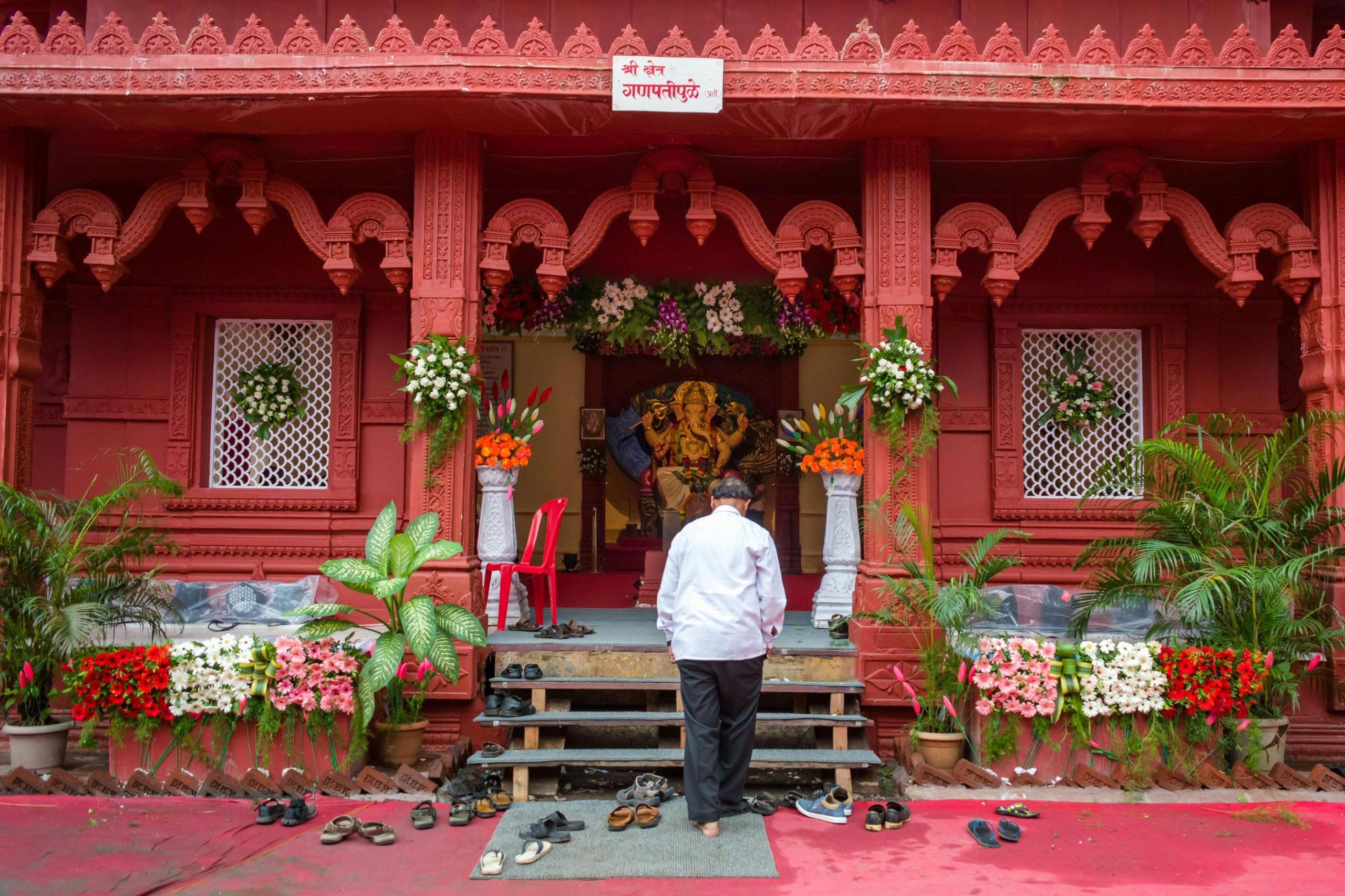 An Indain man dressed in white was standing in front of a bright red coloured temple