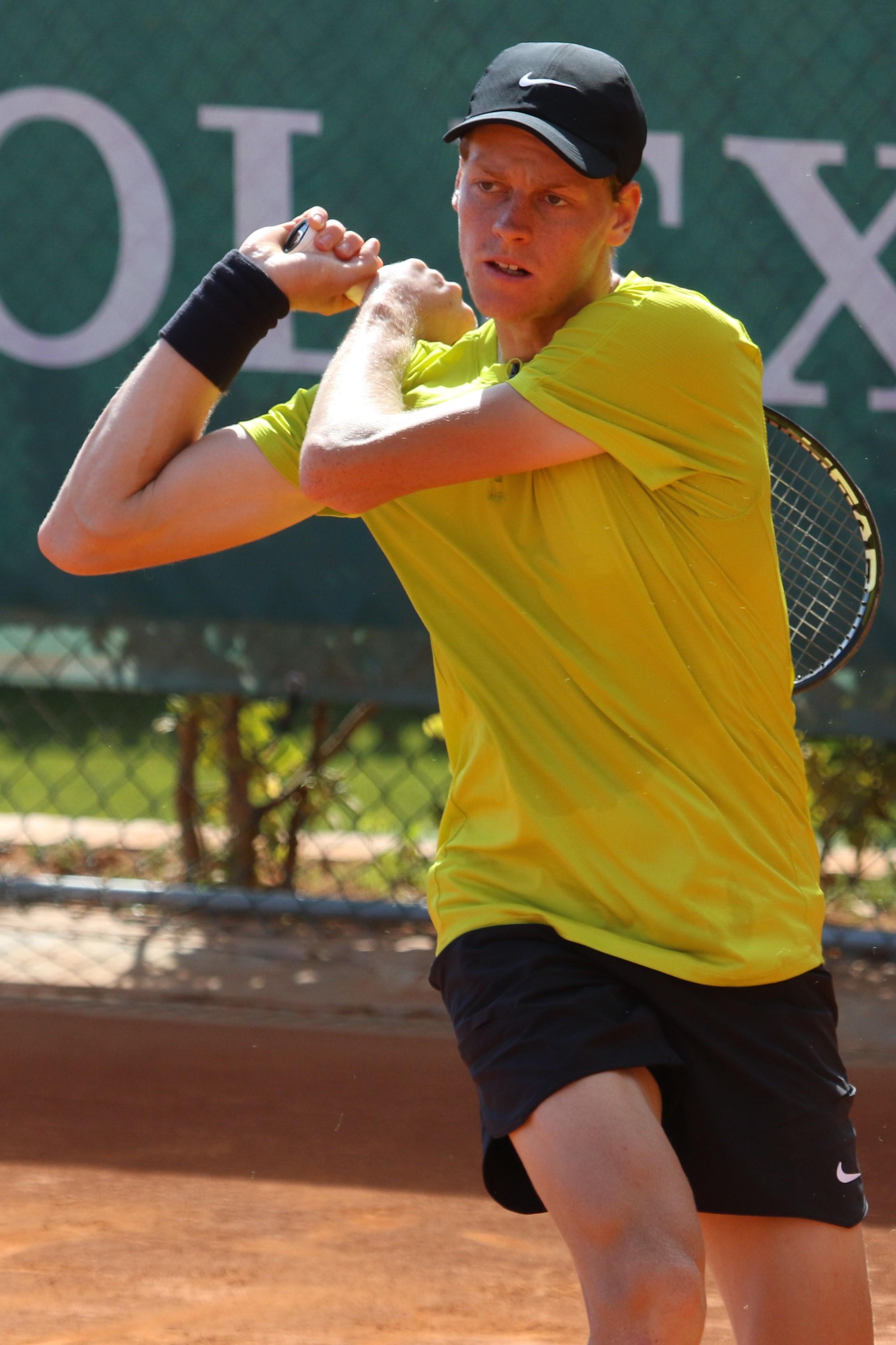 A person wearing a yellow shirt and black shorts, with a black cap on their head, swings a tennis racket on a clay court. 