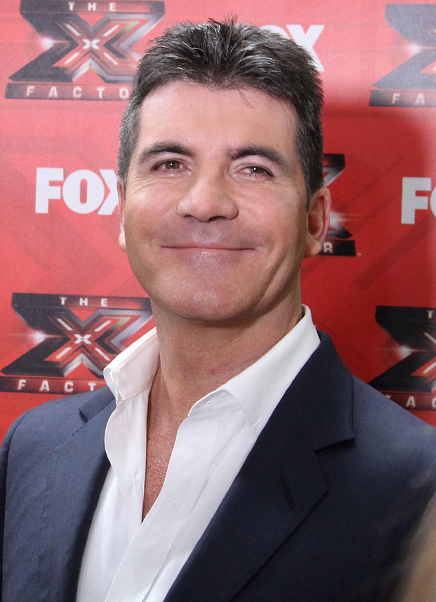 A person wearing a grey blazer over a white button down shirt with an open collar stands and smiles in front of a red, white and black background.