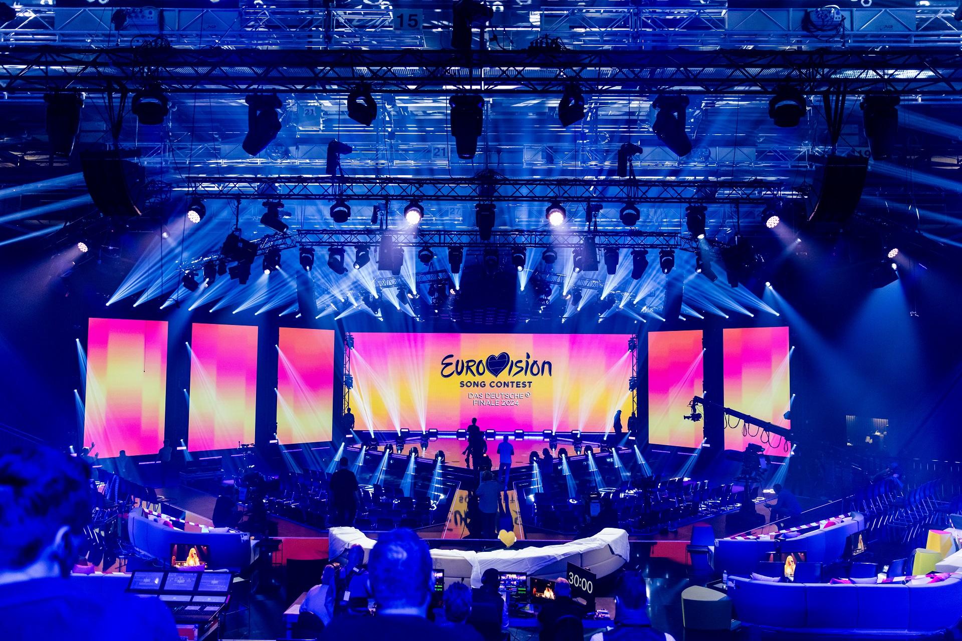 Inside the Eurovision arena, bathed in blue light with the stage backdrop in pink and yellow gradients during set change.