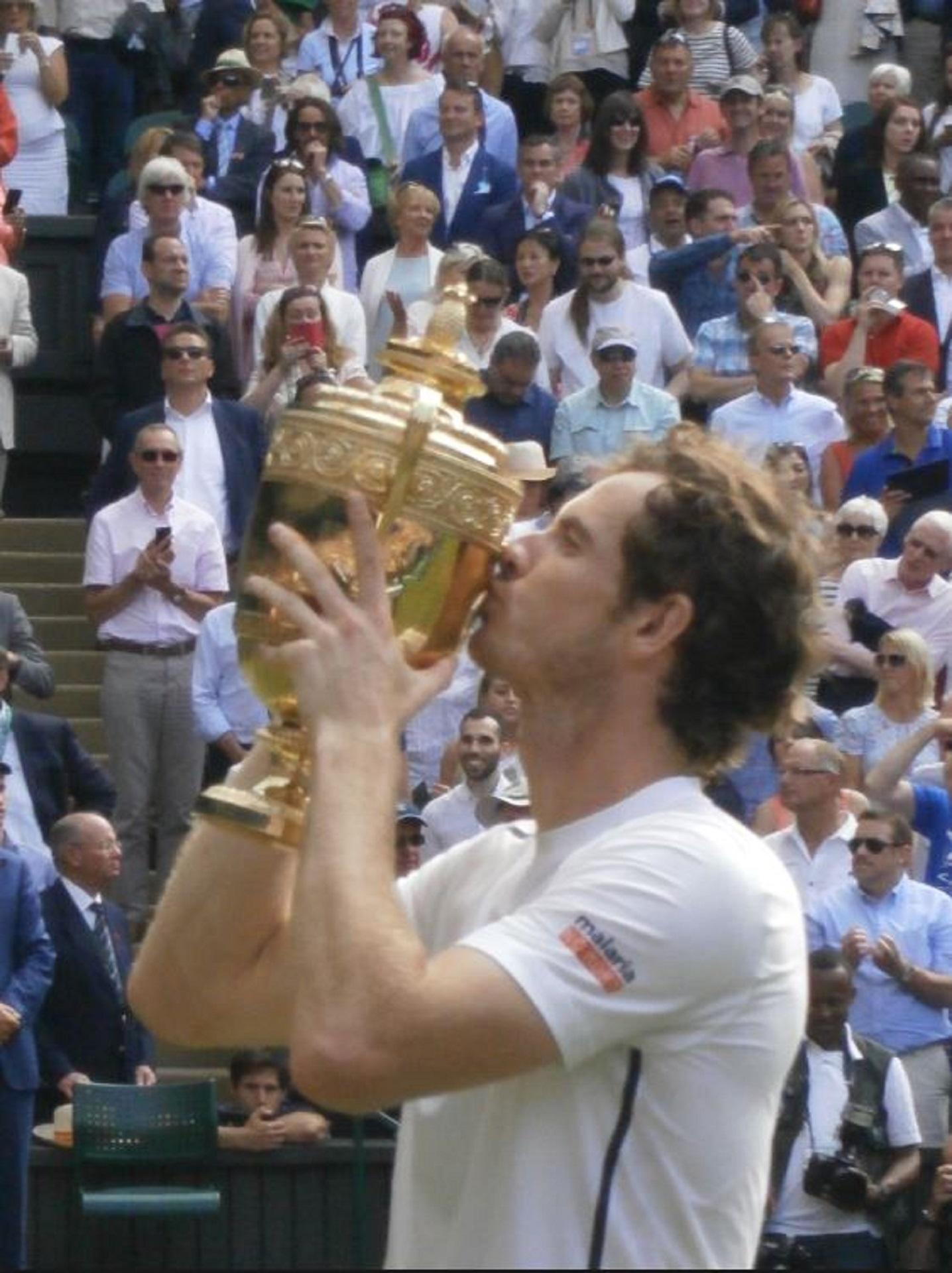 A person wearing a white shirt stands in front of a crowd holding a gold trophy aloft, 