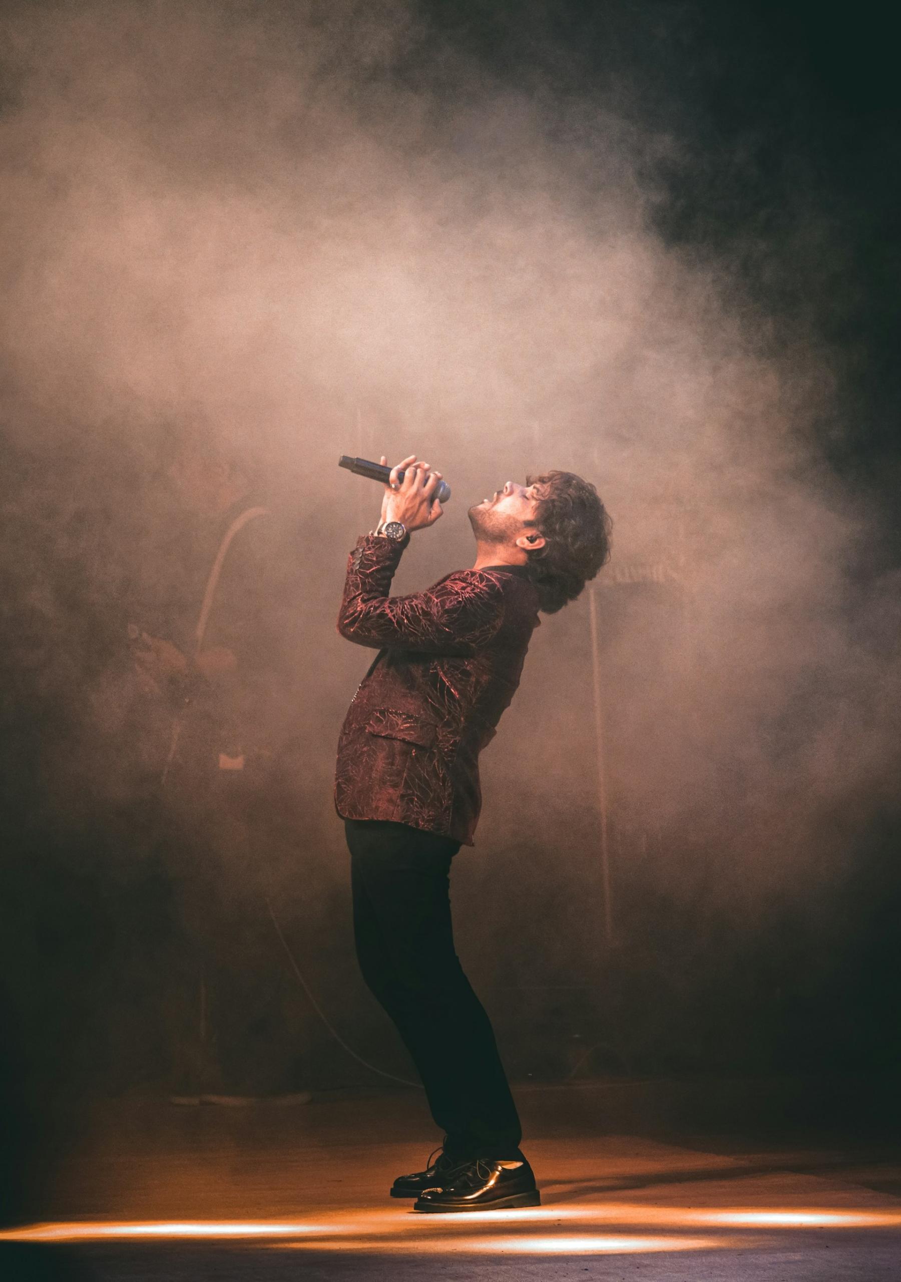 A person wearing a light brown blazer and black slacks holds a microphone to their mouth on stage, spot-lit in a hazy glow.