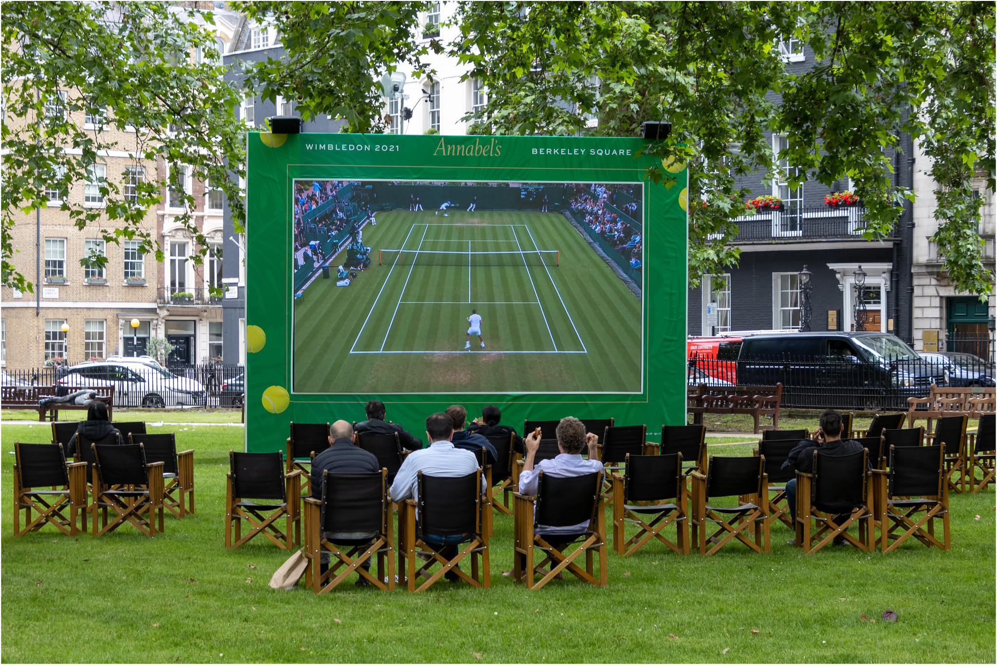 People sitting on a lawn in wooden chairs, watching a tennis match on a big screen.