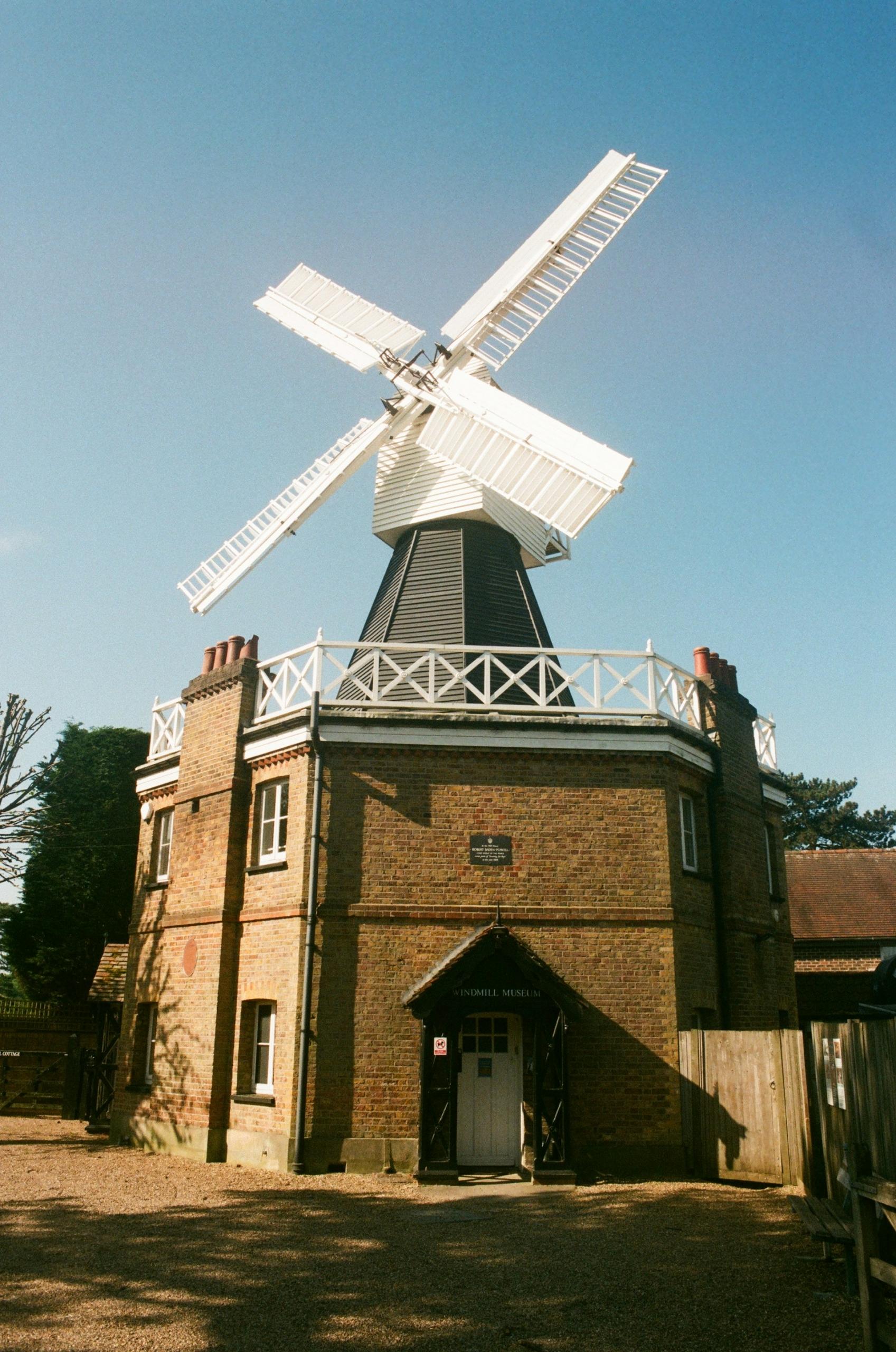 The Wimbledon windmill, a tan brick building with white sails, on a sunny day. 
