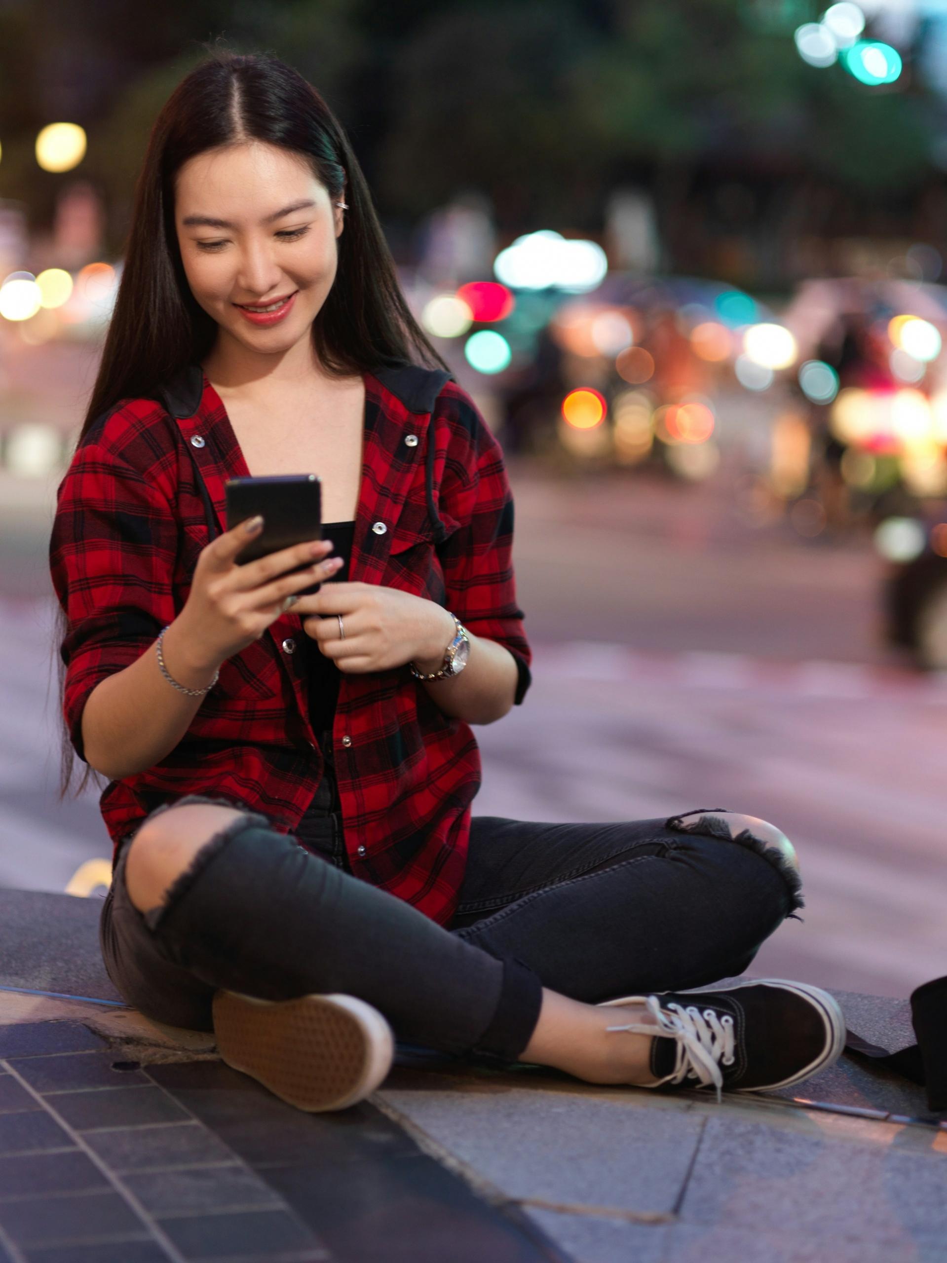 A person in a red and black plaid shirt and torn jeans sits on the sidewalk with a phone in their hand.
